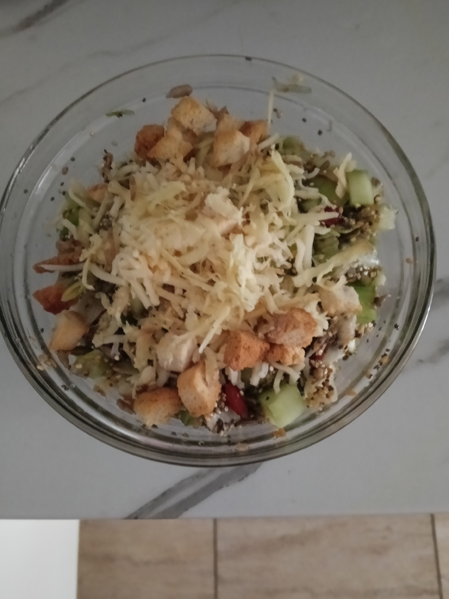 A bowl of salad is shown from an overhead perspective, containing cubed cucumbers, quinoa, shredded cheese, and croutons. The bowl is transparent and sits on a surface with a white and grey marbled pattern. Part of a beige tiled floor is visible in the lower right corner. The location is likely indoors and not necessarily in San Bartolomé de Tirajana, Spain. No people are visible.