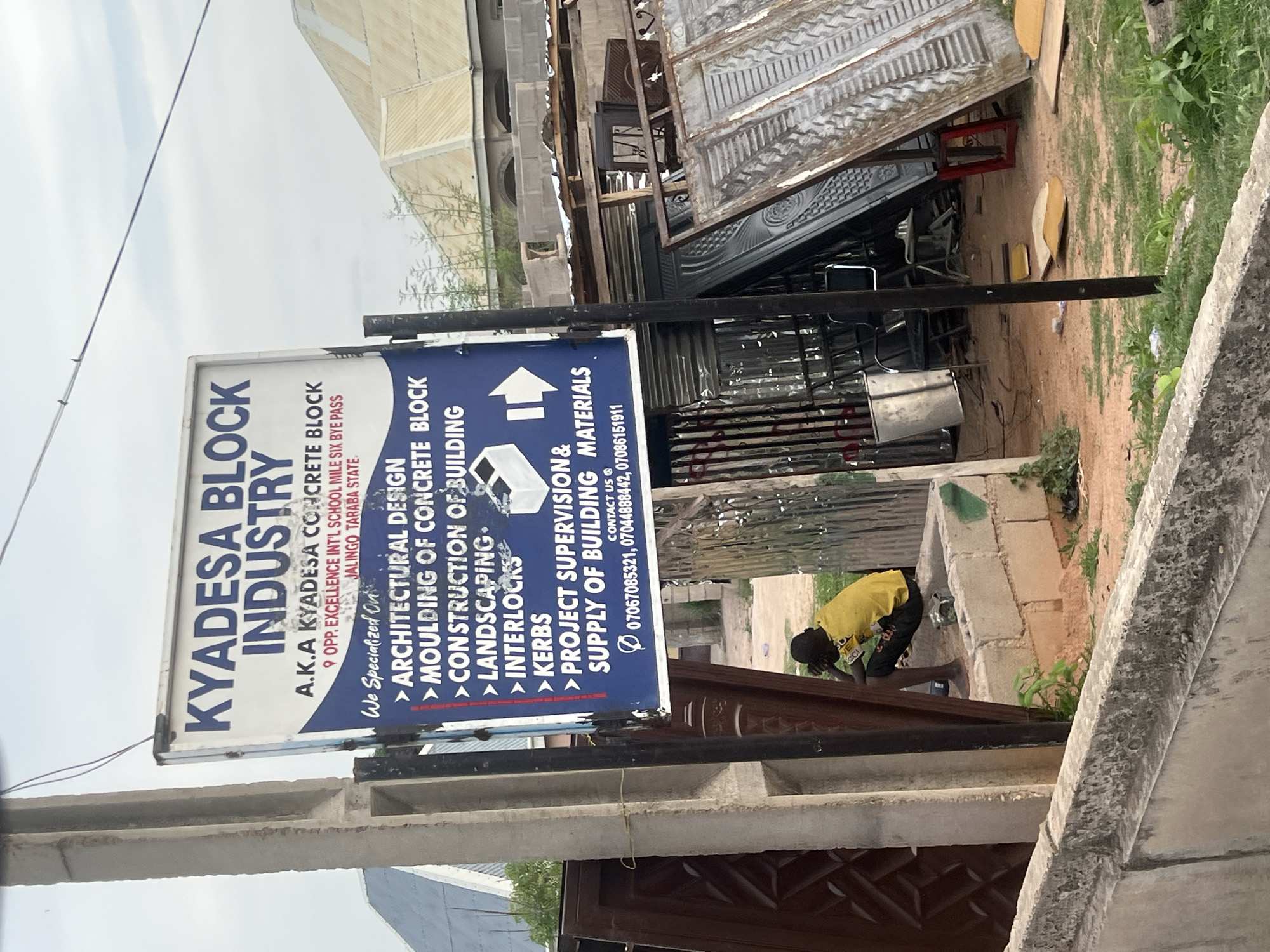 An outdoor, slightly elevated view captures a construction material yard or block industry under an overcast sky. The primary subject is a large, rectangular signboard for "KYADESA BLOCK INDUSTRY," positioned prominently in the mid-ground. The sign, which is mostly white and blue, details the company's services and contact information.
Visible text on the signboard reads: "KYADESA BLOCK INDUSTRY," "A.K.A KYADESA CONCRETE BLOCK," "OPP. EXCELLENCE INT'L SCHOOL MILE SIX BYE PASS," "JALINGO TARABA STATE." It lists specialized services including "ARCHITECTURAL DESIGN," "MOULDING OF CONCRETE BLOCK," "CONSTRUCTION OF BUILDING," "LANDSCAPING," "INTERLOCKS," "KERBS," "PROJECT SUPERVISION & SUPPLY OF BUILDING MATERIALS." Contact numbers "07067085321, 07044888442, 07086151911" are also displayed.
To the right of the signboard, a young person or child wearing a yellow short-sleeved shirt and dark shorts is bent over near a low concrete structure, seemingly engaged in an activity like cleaning or sorting. In the background and around the yard, various building materials are stacked or laid out, including corrugated metal sheets, rebar, and what appear to be finished metal doors or gates. The ground is a mix of dirt and patches of green grass and small plants. Concrete blocks form part of low walls and structures throughout the scene. The overall setting suggests a bustling industrial or commercial area focused on construction, likely in Howayi, Jalingo, Taraba State, Nigeria, as indicated by the signboard. The time of day appears to be daytime with soft, diffused lighting.