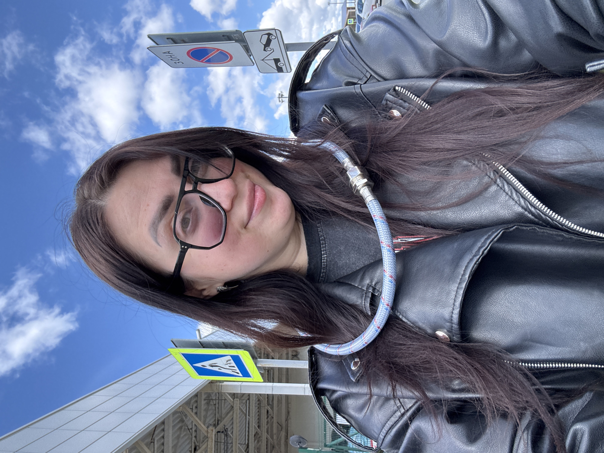An outdoor, slightly rotated selfie captures a person from the chest up, looking directly at the camera under a bright blue sky with scattered white clouds. The subject is a person with long, dark brown hair, wearing black-framed glasses and a black leather jacket over a dark top. A light blue and white braided rope or cable, featuring silver metal connectors at the ends, is draped around their neck and shoulder.
In the background, against the clear sky, several road signs are visible. On the upper left, an inverted circular sign with a red border, blue interior, and a red diagonal line signifies "No Stopping" or "No Parking," with the Cyrillic text "3OHA" (Zone) clearly readable below it. Next to it is a rectangular sign depicting a car being towed, indicating a towing zone. Lower down and to the left, a blue square sign with a white triangle and a walking pedestrian symbol suggests a pedestrian crossing. Behind the subject, to the left, parts of a modern building with a grey, grid-like facade are visible. The person appears to be taking the photo in an urban or semi-urban environment on a clear, sunny day.