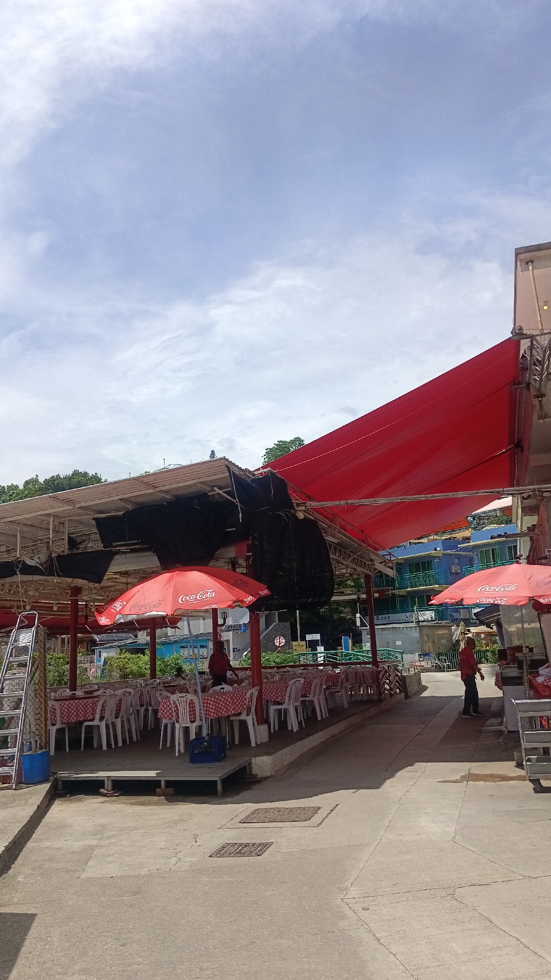 The image depicts an outdoor dining area, likely a restaurant or food stall, situated in Hong Kong, China. The scene is characterized by red and white checkered tablecloths covering numerous small tables, each surrounded by white plastic chairs. Overhead, a large red tarp serves as a canopy, providing shade. Red umbrellas, some emblazoned with the "Coca-Cola" logo, are also visible, further protecting the seating area. In the background, multi-story buildings with blue and green facades are present, suggesting an urban or semi-urban environment. Two individuals in red shirts are visible, one appearing to be working in the establishment and the other walking away from the camera. The ground is paved with concrete. The overall impression is a casual, open-air dining setting on a bright day.