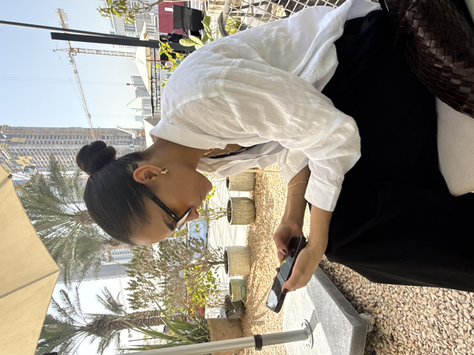 A young adult female is depicted in profile, seated outdoors in a sunny urban environment in Dubai, United Arab Emirates, likely on a cafe patio. She has dark hair styled in a high bun and wears black-framed sunglasses with amber-tinted lenses, small gold hoop earrings, a white collared long-sleeved shirt, black trousers, and a thin gold necklace and bracelet. Her fingernails have dark polish. She is focused on a dark-colored smartphone, holding it with both hands and her thumbs positioned for interaction.
The immediate surroundings feature a light-colored outdoor umbrella partially visible at the top left, providing shade. The ground is covered with light-colored gravel, and several potted plants, including palm trees and smaller shrubs, are arranged around the seating area. A white metal chair frame is partially visible to her right, and a woven dark brown bag rests beside her. In the background, multiple modern high-rise buildings, some appearing to be under construction, dominate the skyline. At least two large construction cranes are prominent, indicative of ongoing development in the city.