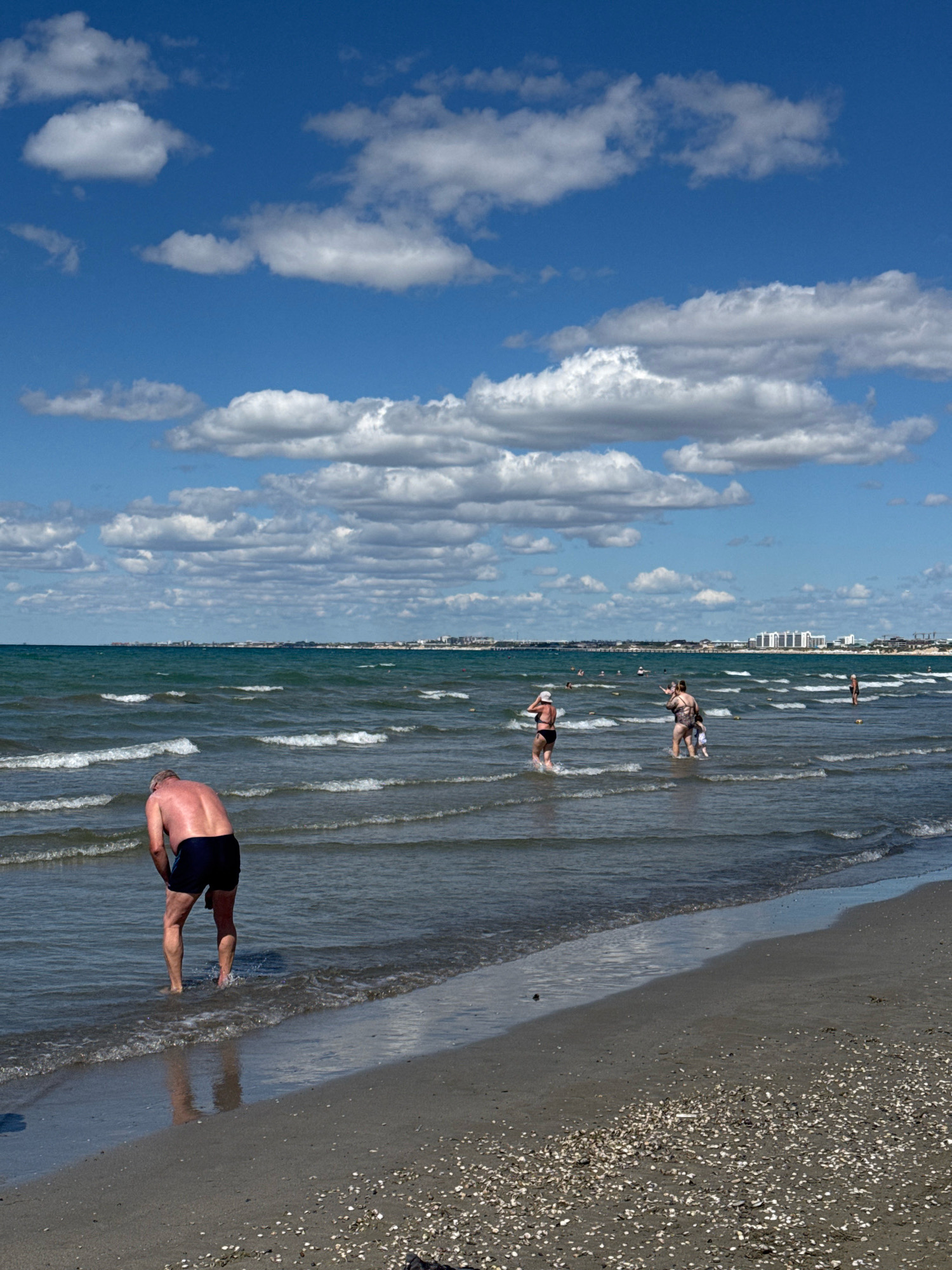 A wide shot captures a beach scene on a bright, sunny day with fluffy white clouds scattered across a vivid blue sky. The ocean is a deep green, with gentle waves rolling towards the shore. Several people are visible in the water and on the beach. In the foreground, a man with tanned skin and dark shorts stands facing away from the camera, with his back to the viewer, as he enters the water. Further out in the sea, a group of people are wading and interacting. In the background, a distant coastline is visible, dotted with buildings that suggest a city or town. The beach itself is composed of dark sand speckled with shells, and the water's edge is lined with a thin layer of foam from the waves.