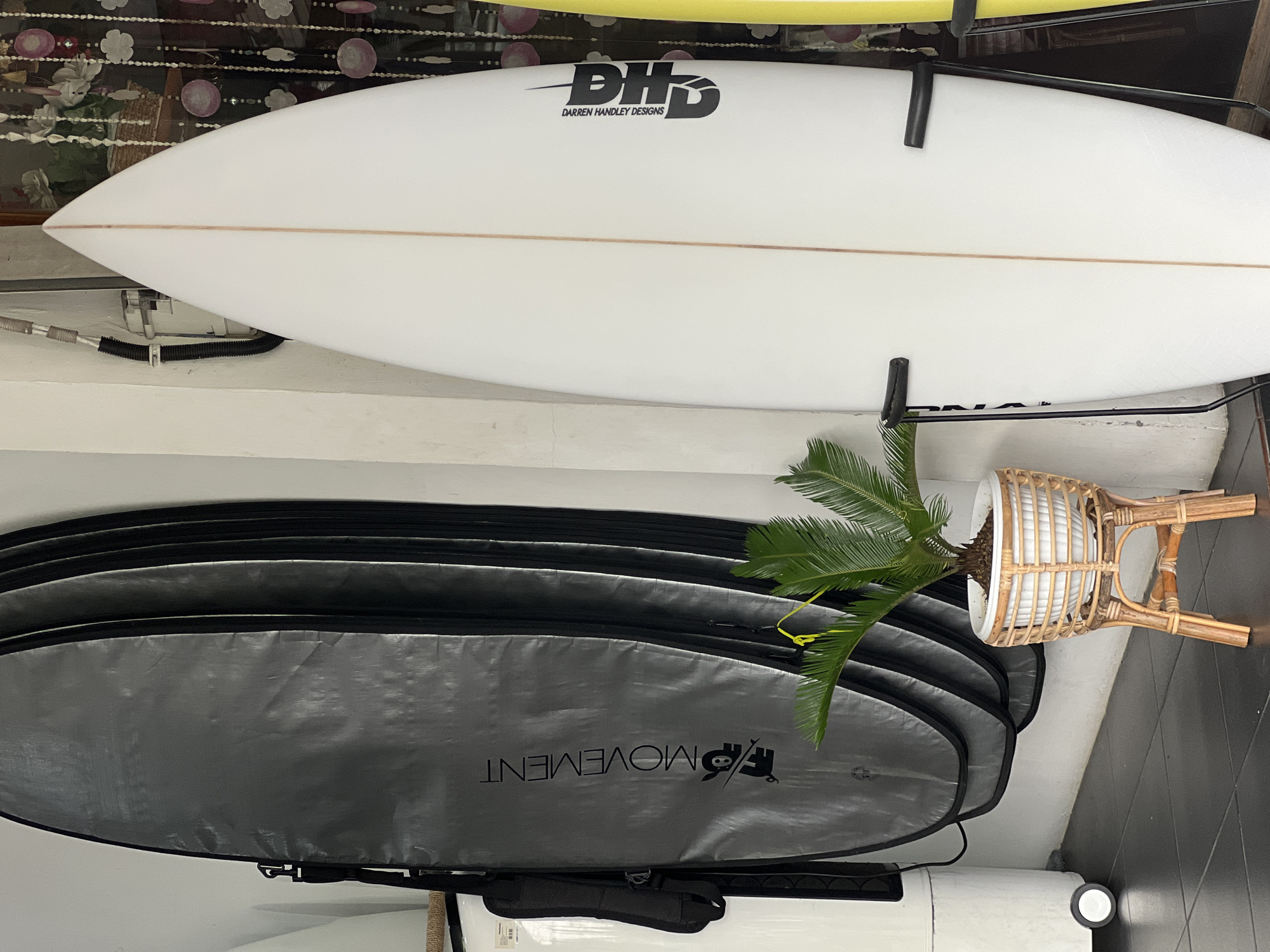 This is a medium shot, taken at eye level, of a collection of surfboards in what appears to be a shop or storage area in Kuta, Indonesia. The background is a white wall, with some decorative beaded curtains and a basket with flowers visible on the far right.
In the foreground and center, a white surfboard with a single brown stringer is propped upright in a black metal stand. The surfboard has black lettering on the right side that reads "PHD DARREN HANDLEY DESIGNS." To the left of the white surfboard, several grey surfboard bags are stacked vertically, with one bag prominently displaying a black logo and the word "MOVEMENT" in black text. A small potted cycad plant with green fronds sits in a woven rattan planter on a wooden stand to the left of the white surfboard.
To the far left, a white cylindrical object with wheels is partially visible, suggesting it might be a piece of equipment or storage. The floor is dark grey tiled. The lighting is even and appears to be natural or well-diffused artificial light. No people are visible in the image. The overall impression is of a well-organized space dedicated to surfing equipment.
