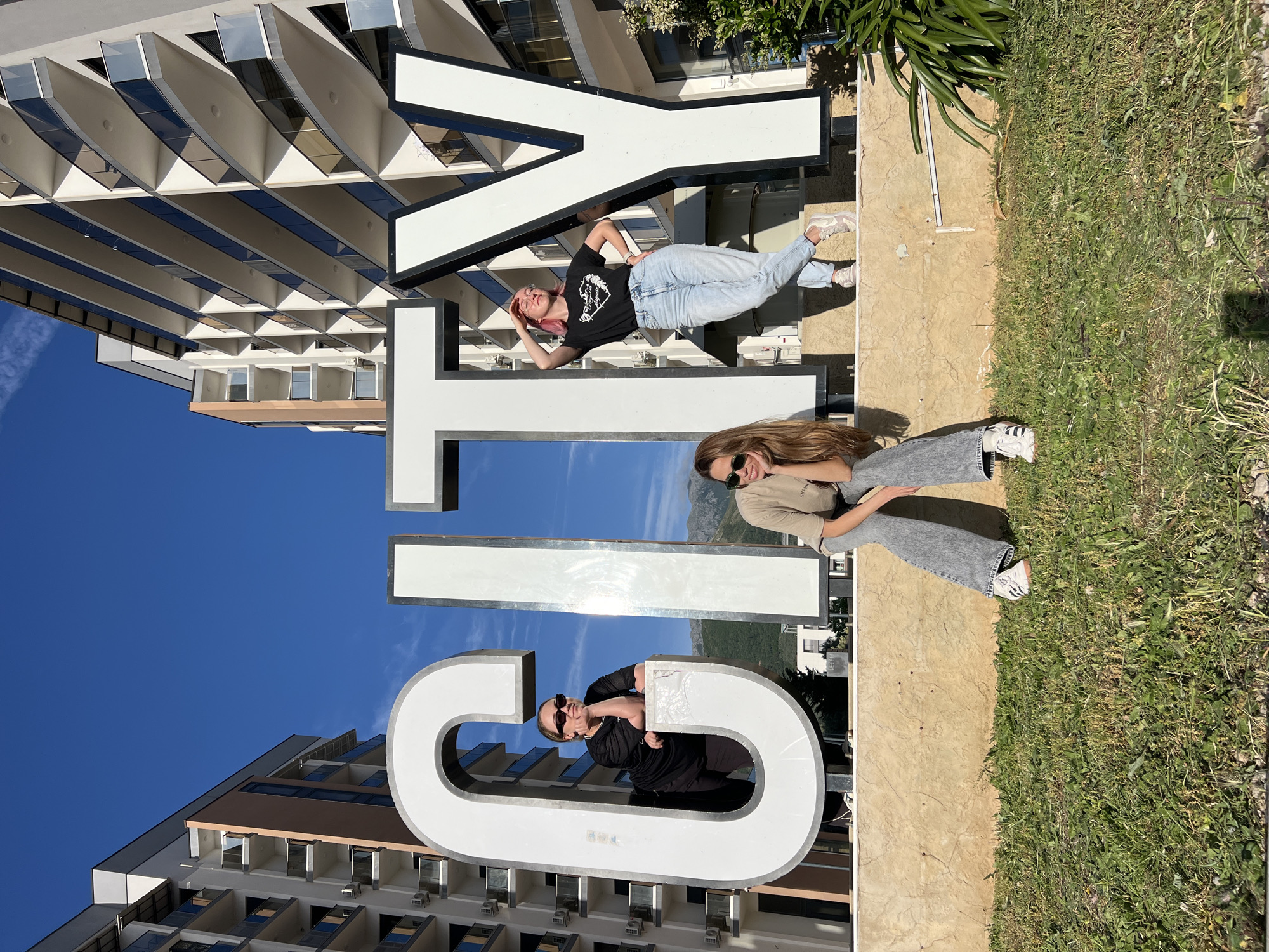 Three people are interacting with a large sculpture that spells "Bar" in the city of Bar, Montenegro. The letters are large, white, and freestanding. A building with mirrored sunshades is visible behind the letter 'Y'. To the right, grass borders the sculpture's base. A woman with pink hair lies across the 'Y', another sits on the 'T', and a third poses within the 'B'. Each is looking towards the camera. The sky is clear and blue.