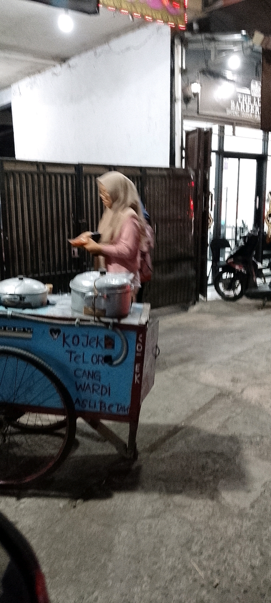 In Karangsatria, Indonesia, a woman wearing a light-colored hijab and a pink long-sleeved shirt is standing next to a food cart. The cart is blue with red trim and has writing on its side, including "KOJEK TELOR," "CANG WARDI," and "ASLI BETAWI." Two silver pots with lids are on top of the cart. The woman appears to be holding a small object in her hands. Behind the cart, a metal fence is visible, and further back, a motorcycle is parked. The scene takes place on a paved surface, possibly a street or sidewalk, with shadows cast by the cart and other elements. A sign for "THE LUX BARBER" is visible in the background.