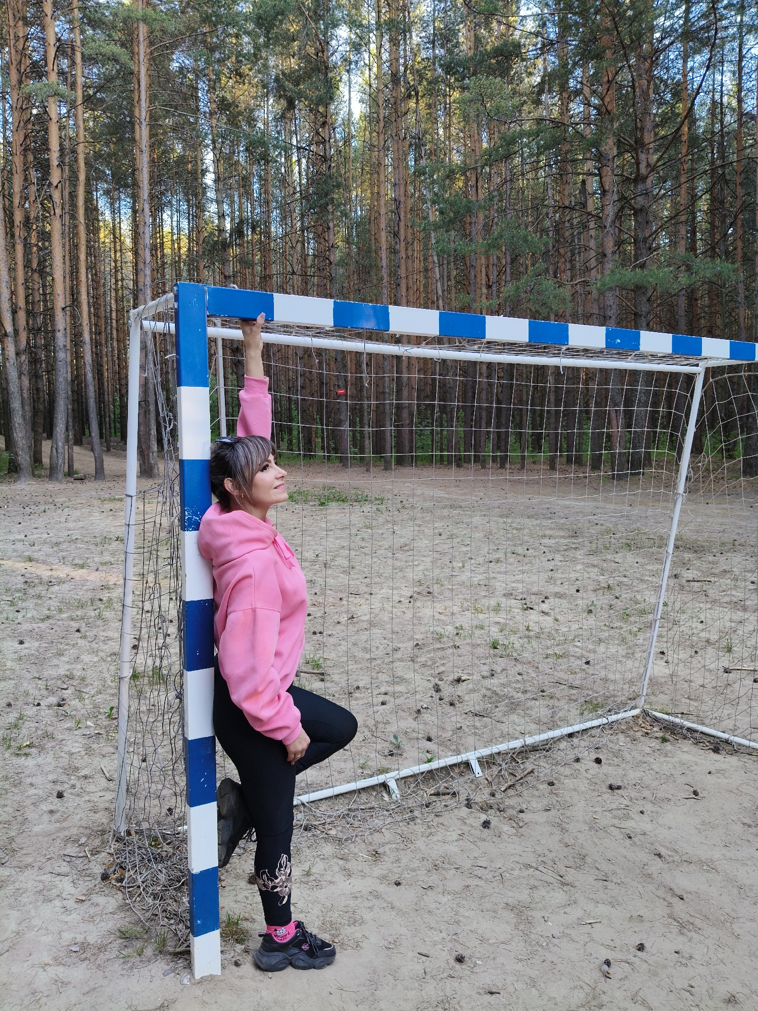 A woman is standing next to a soccer goal in a sandy area in Chaikovsky, Russia. The woman has dark hair, bangs, and is wearing a pink hooded sweatshirt and black leggings. She is leaning against the goalpost, with her left leg bent at the knee and foot resting on the post. Her left arm is raised, and she is holding onto the top of the goalpost. She is looking upwards, away from the camera. The soccer goal is white with blue and white stripes on the top bar and left post. It is surrounded by a net. The ground is sandy, with scattered leaves and small pine cones. In the background, there is a dense forest of tall, thin pine trees.
