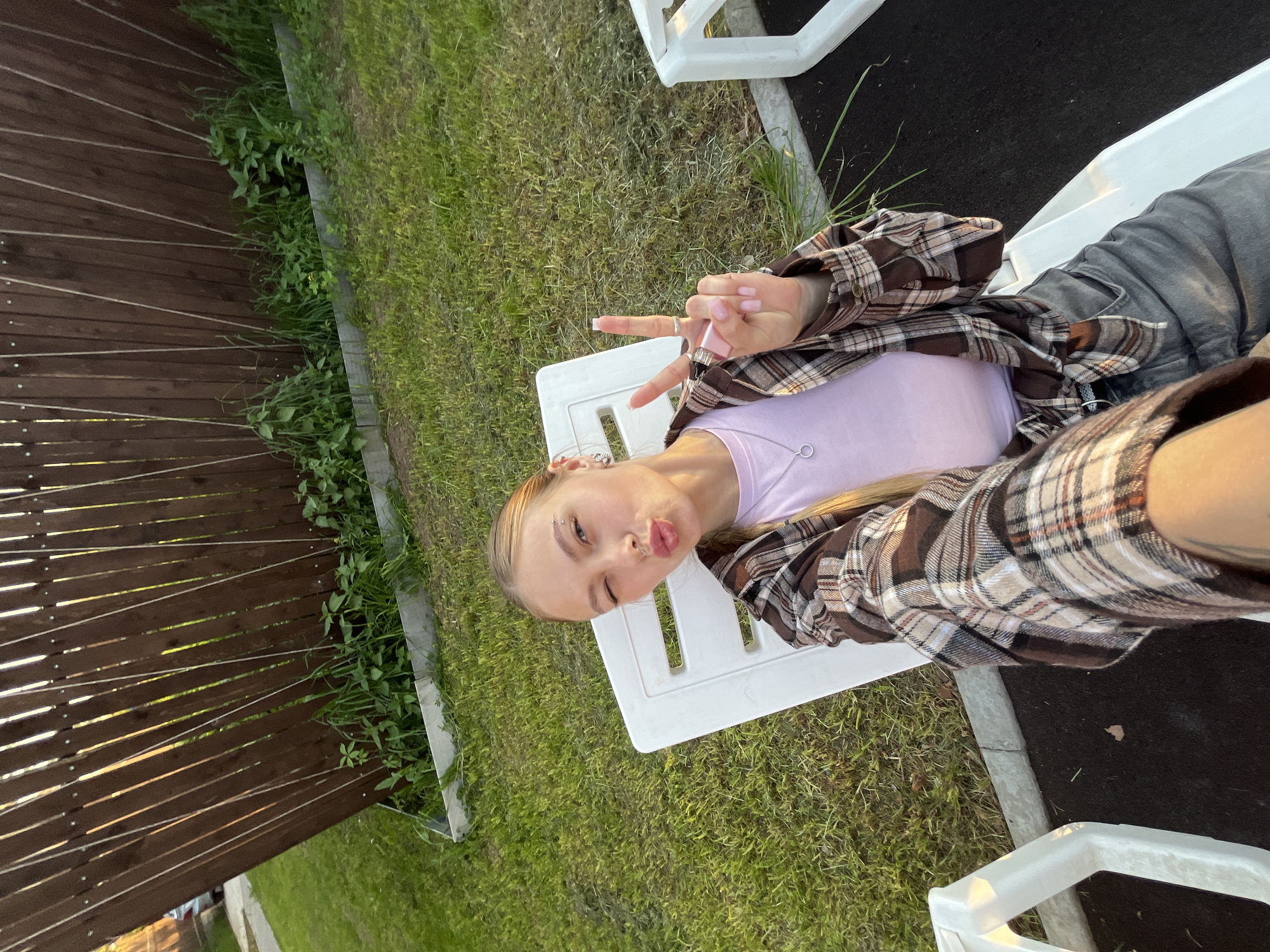 A young woman is lying on a white plastic lounge chair outdoors on a sunny day. She is wearing a light purple t-shirt and a brown and beige plaid shirt. She has long blonde hair and is wearing gray jeans. She is making a peace sign with her right hand and puckering her lips. The chair is situated on a lawn next to a wooden fence with horizontal slats. In the background, there is a black paved surface. The location is in Chaikovsky, Russia.
