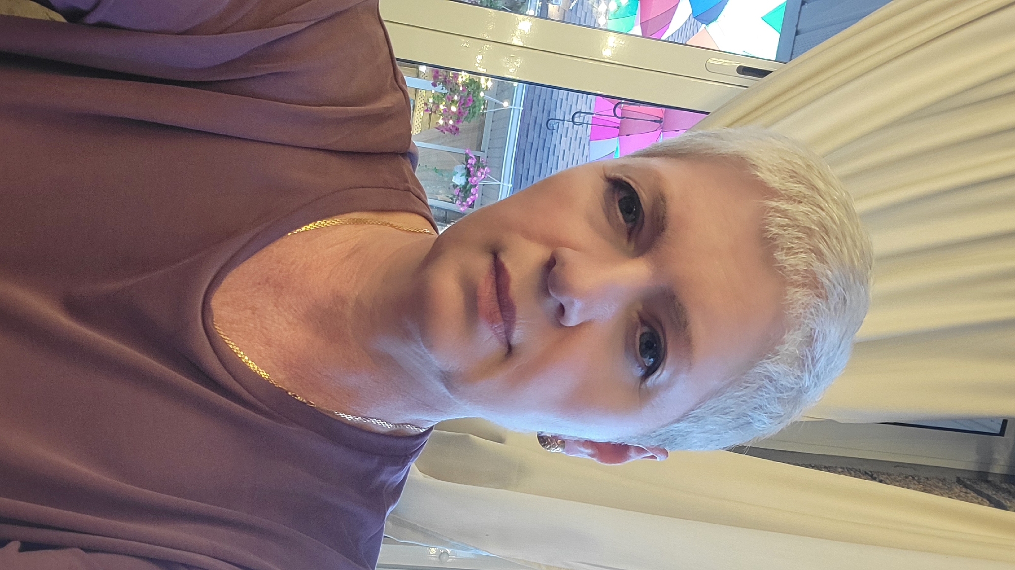 A woman with short, white hair and blue eyes is photographed from the chest up. She is wearing a dark purple, V-neck t-shirt and a gold chain necklace. She has gold hoop earrings in her ears. Behind her, to the left, is a window with curtains. Through the window, colorful umbrellas are visible, as well as hanging flower baskets with pink and purple flowers. The background appears to be outdoors, possibly a courtyard or patio area. The lighting is soft and natural. The woman's expression is neutral.