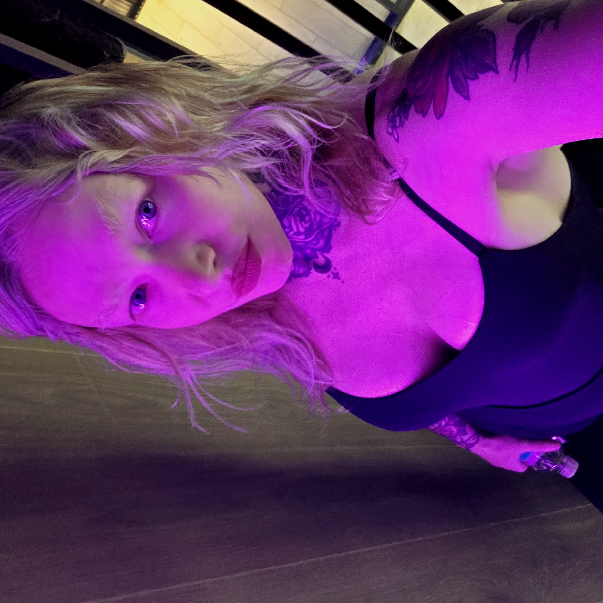 A woman with blonde, wavy hair and light eyes poses in a low-angle selfie. The scene is predominantly lit with a vibrant pink light. She is wearing a black tank top, revealing a tattoo on her upper arm. Another tattoo is visible on her neck. In her hand, she holds what appears to be a water bottle. The background is blurred, suggesting an indoor setting, but no further details allow one to identify a precise location within Saint Petersburg, Russia.