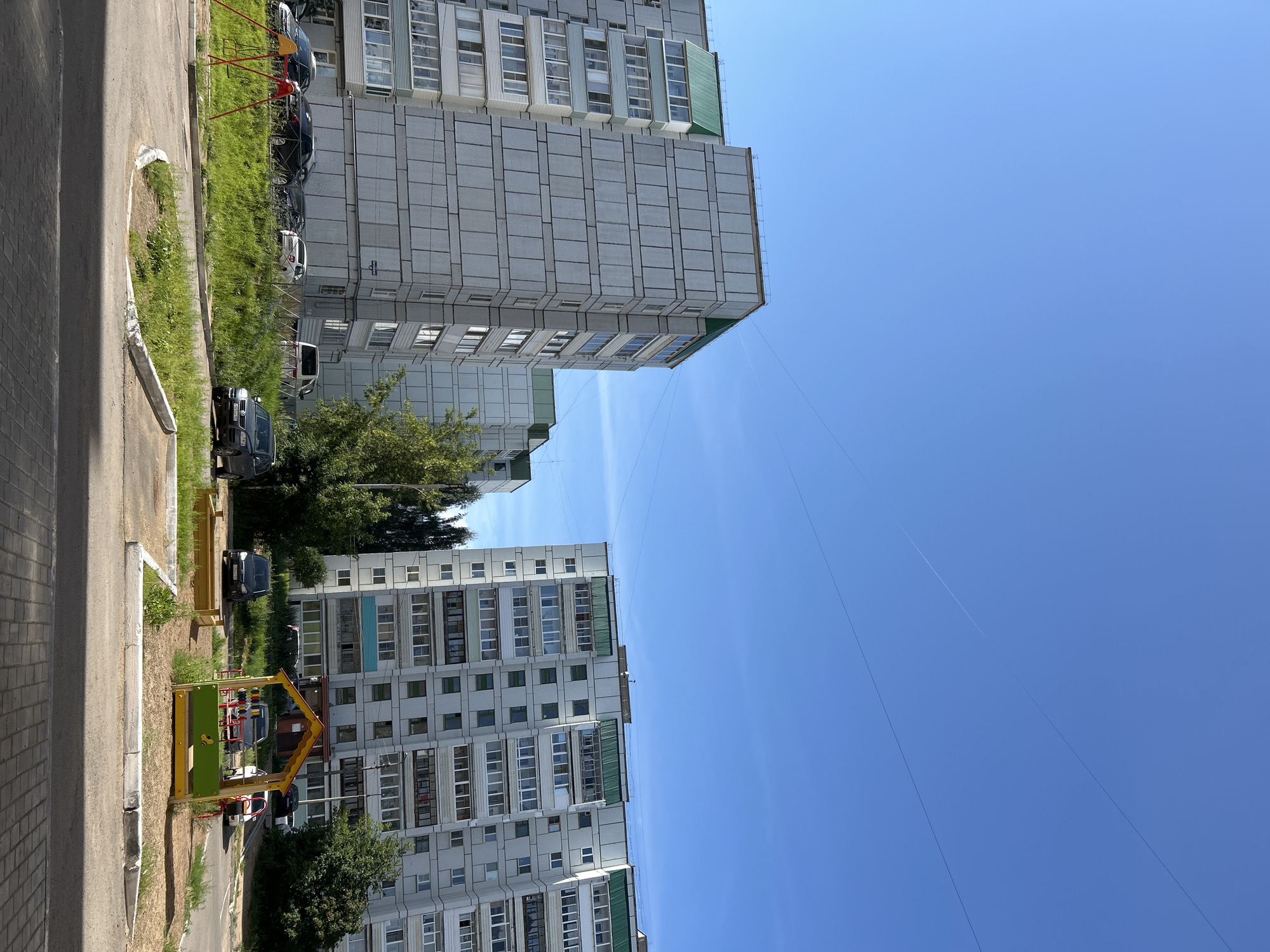 The image depicts an outdoor scene in the city of Chaikovsky, Russia, on a clear, sunny day. The foreground shows a paved area with a curved concrete curb, leading to a patch of grass with a small sandbox and a colorful children's playhouse with a yellow roof and green sides. A swingset with red supports is also visible in the grassy area. Beyond the playground, a line of parked cars, including a dark SUV, a white sedan, and several other vehicles, are seen in front of a row of apartment buildings. The buildings are multi-story structures with gray facades and white-framed windows, some with green awnings or balconies. Trees and foliage are interspersed between the buildings and behind the cars. The sky above is a clear, vibrant blue with faint lines, possibly from aircraft contrails.