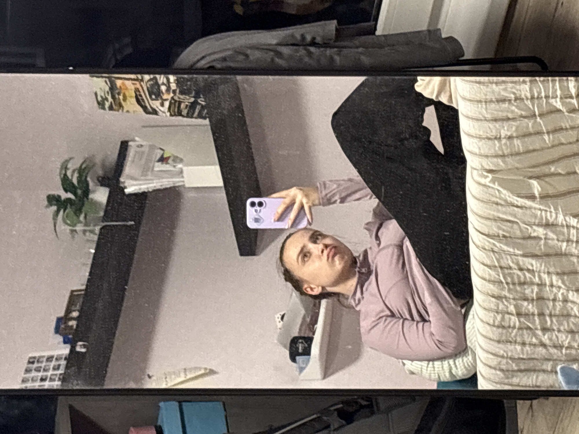 This mirror selfie captures a young woman indoors in what appears to be a bedroom or living area in Прикамский, Russia. The image is oriented sideways, showing the woman's reflection as she holds a light purple smartphone, taking the picture. She has light skin, dark hair pulled back, and is wearing a light purple long-sleeved top, possibly a hoodie, with a slightly pouting or serious expression.
She is seated or reclining on a soft surface, likely a bed, covered with light-colored bedding featuring subtle stripes. In the background reflection, a dark, multi-tiered shelf unit is visible against a light wall. On the shelves, various items can be seen, including a small green potted plant, stacks of papers or books, and what might be small framed pictures or decorative tiles. A white storage bin is also present. Above the shelves, a dark gray garment or blanket is draped. The mirror itself has a dark border. No discernable text is visible.