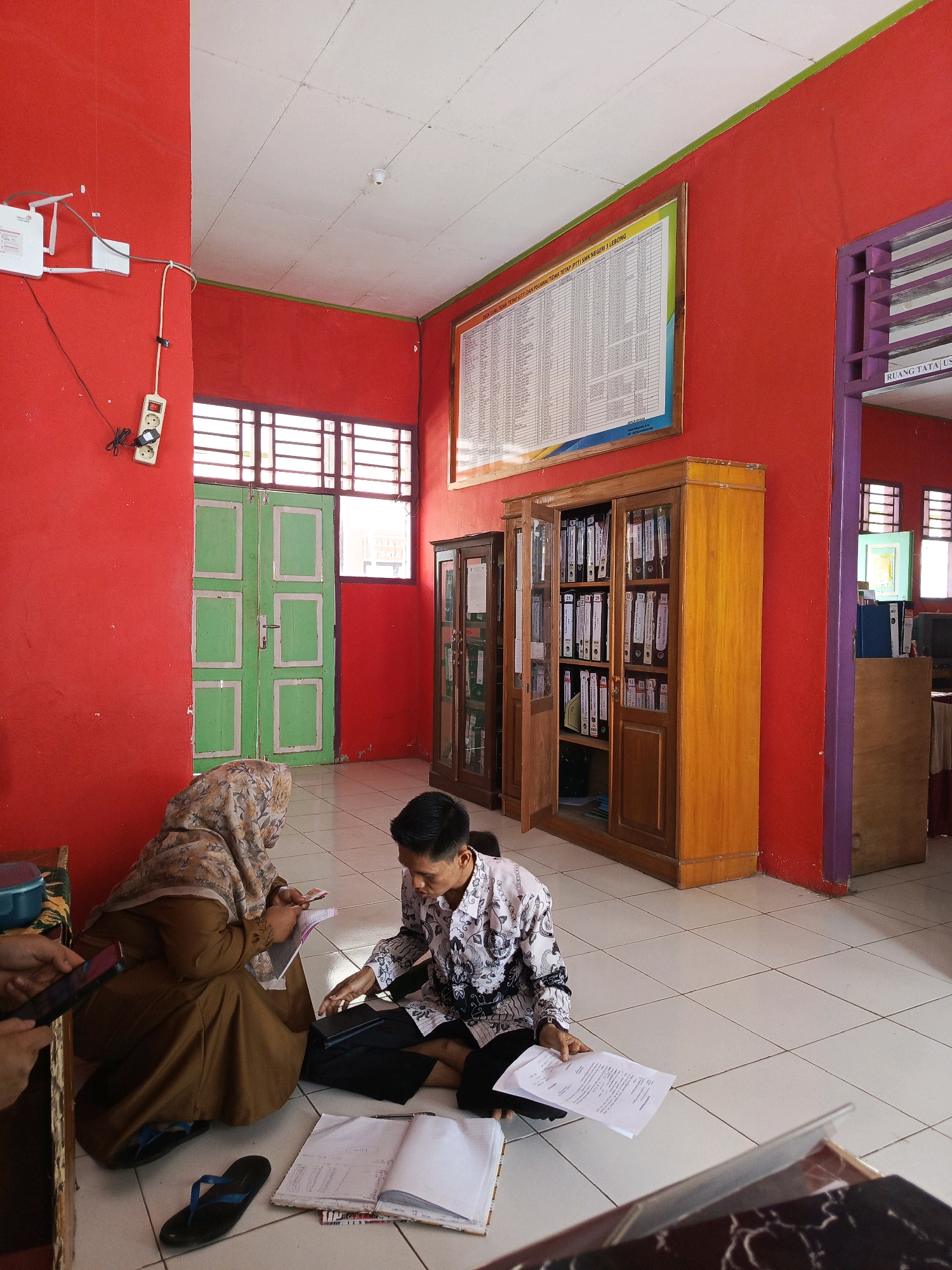 The image depicts an indoor scene within what appears to be a classroom or office space in Tik Jeniak, Indonesia. The walls are painted a vibrant red, and the ceiling is white. The floor is tiled in a grid pattern. Two individuals are present, both seated on the floor. One, a woman wearing a hijab and a brown garment, is looking down at a book or papers in her hands. The other, a man wearing a patterned shirt, is holding a document and looking towards the floor. Several books and papers are scattered around them on the floor. In the background, there is a wooden cabinet filled with binders, a green door, and a large chart or notice displayed on the wall. Electrical outlets and wiring are visible on the red wall.