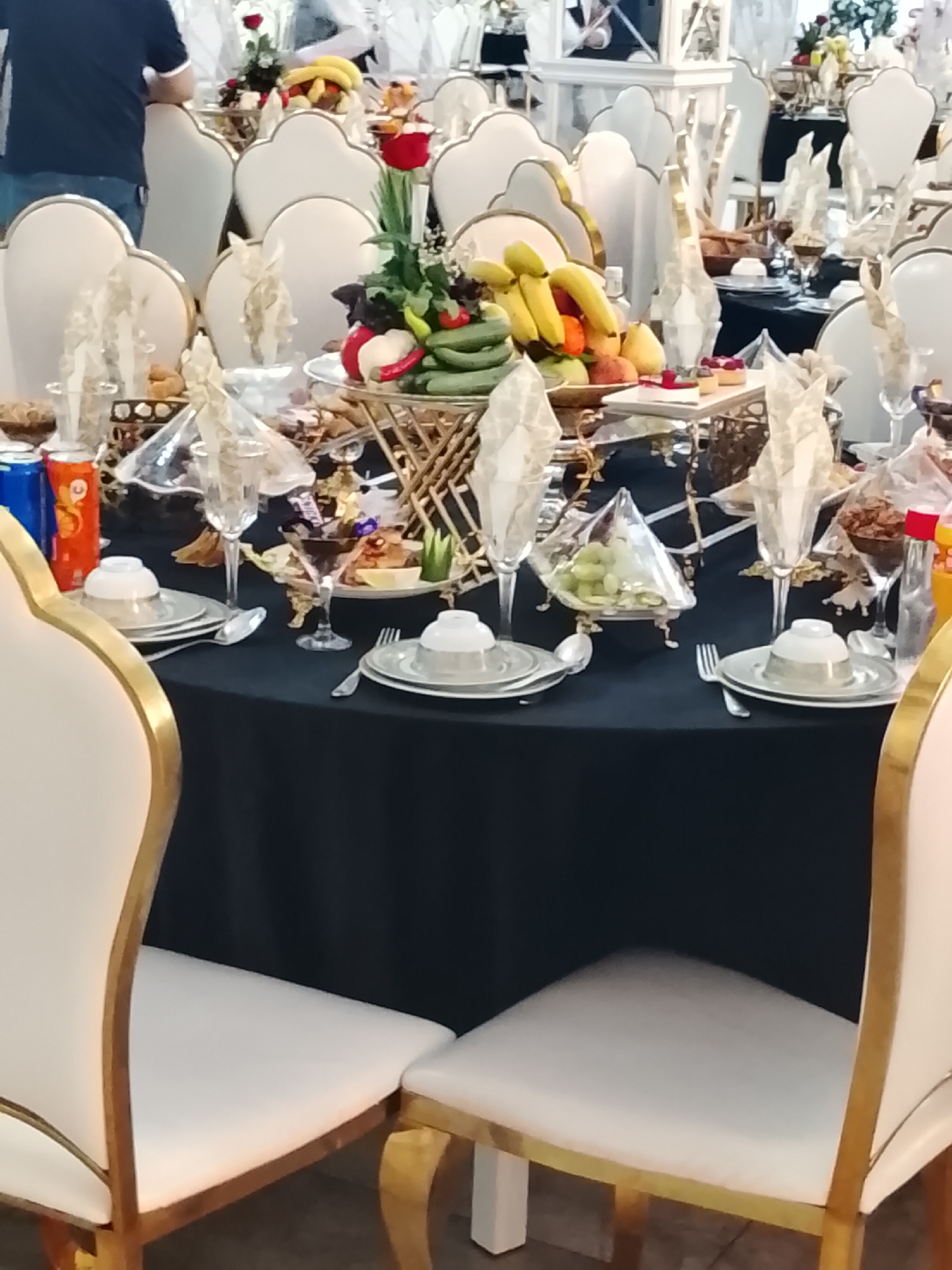 The image depicts a formal dining table set for a celebration or event, likely a wedding or large gathering, in Urgench, Uzbekistan. The table is draped in a black tablecloth and adorned with white plates, silver cutlery, and elegant glassware. A centerpiece of fruits, including bananas and what appear to be pomegranates and cucumbers, is prominently displayed. Several chairs with white upholstery and ornate gold trim surround the table. In the background, other tables and chairs are visible, suggesting a larger event space. The scene conveys a sense of opulence and careful preparation for guests.