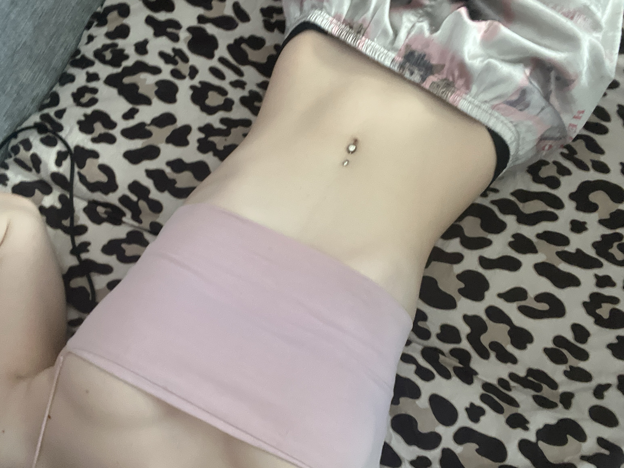The image depicts a close-up view of a person's torso lying on a bed with a leopard-print sheet. The individual is wearing a light pink, form-fitting top and patterned pajamas. A navel piercing is visible. The image does not contain information to determine if it was taken in Saint Petersburg, Russia.