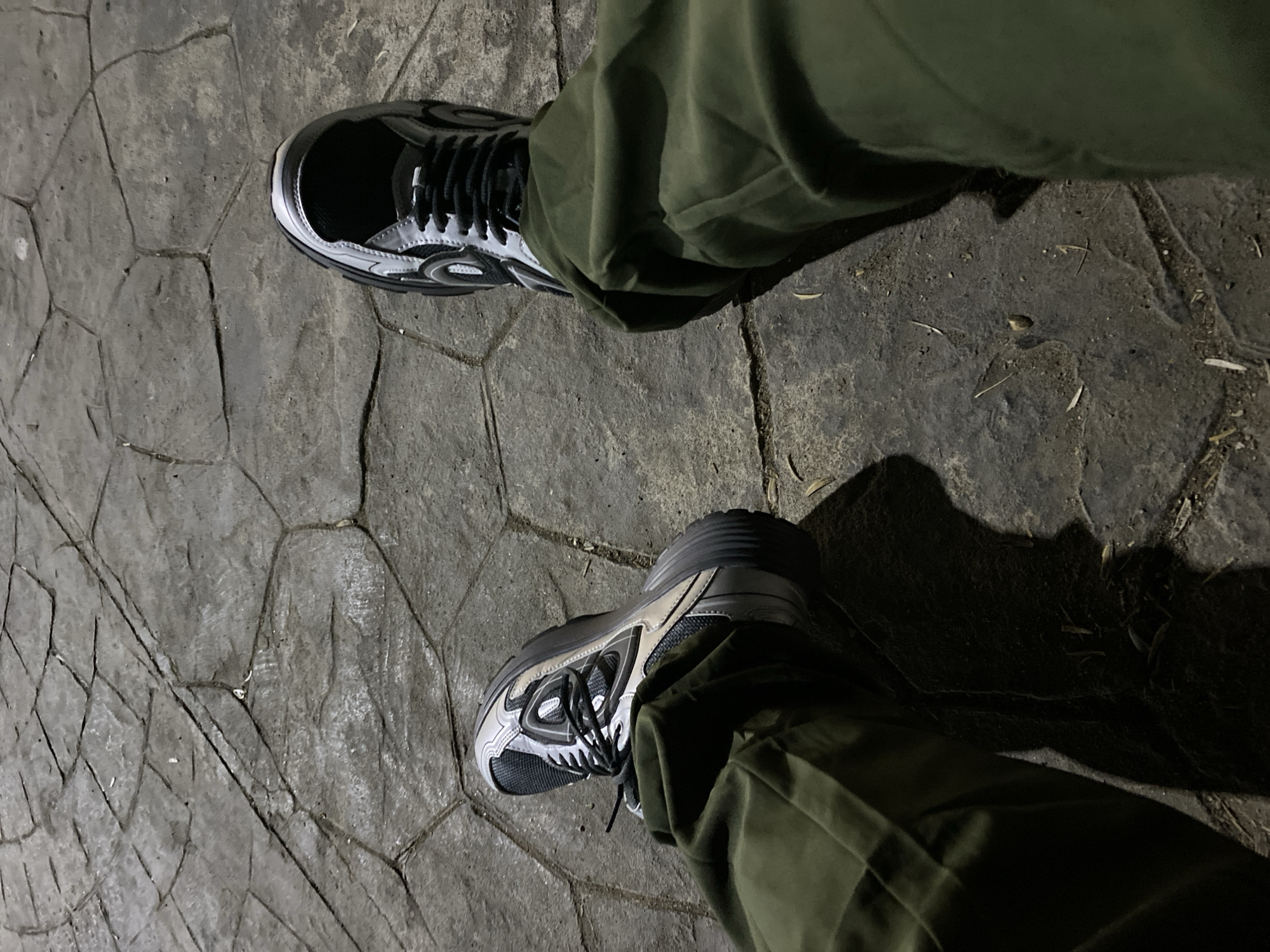 The image shows a person's legs and feet wearing olive green pants and grey and black athletic shoes. The shoes have a chunky sole and intricate detailing. They are standing on a textured, grey stone surface that appears to be paving stones arranged in an irregular pattern. The lighting suggests it is either nighttime or a dimly lit indoor space, casting shadows from the person's legs onto the ground. The environment is consistent with an outdoor urban setting, possibly a public walkway or plaza. There is no indication of the specific location being in Tipaza, Algeria.