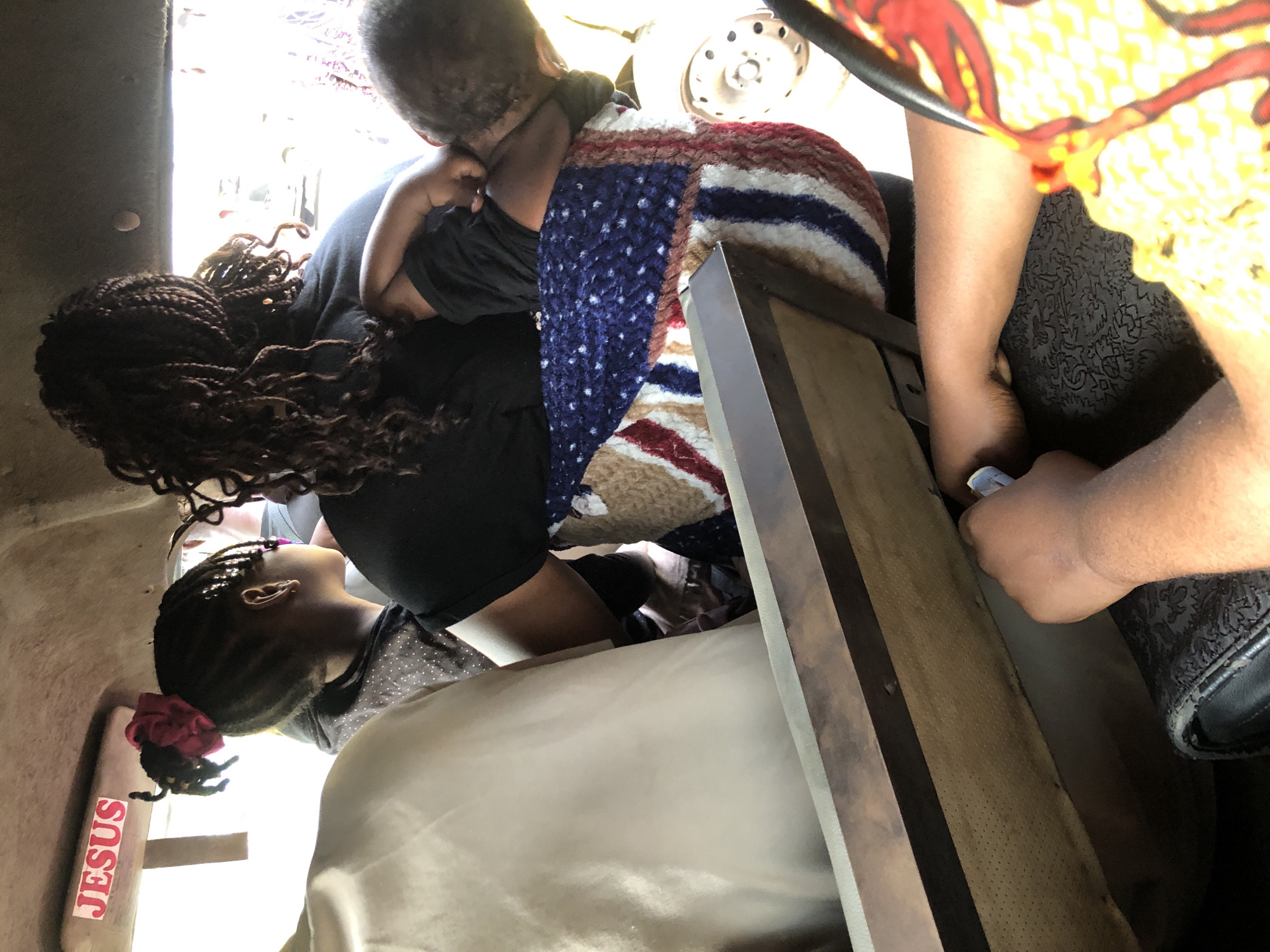 The image captures the interior of a vehicle, likely a bus or taxi, in Ikeja, Nigeria. A woman with braided hair is seen from behind, holding a baby wrapped in a blanket with a patriotic American flag design. To her left, a young girl with a red bow in her hair looks to the side. The vehicle's interior is somewhat cramped, with the heads of other passengers visible. A sticker with the word "JESUS" is affixed to the sun visor. The overall impression is a snapshot of everyday life within a crowded public transport setting in the bustling city.