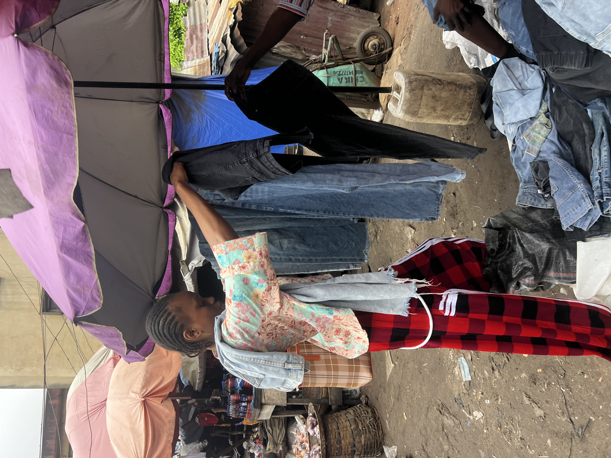 A woman with braided hair, wearing a colorful floral-patterned shirt and red and black checkered pants, stands in an outdoor market stall. She is holding up a pair of black pants and a pair of blue jeans. To her right, another person's hand is visible, holding black pants. Various other articles of clothing, including more jeans, are piled on the ground around them. The stall is covered by a large purple umbrella, and there are other umbrellas and market structures visible in the background. The ground appears to be dirt and is littered with debris. The scene is set in the city of Nkpor, Nigeria.