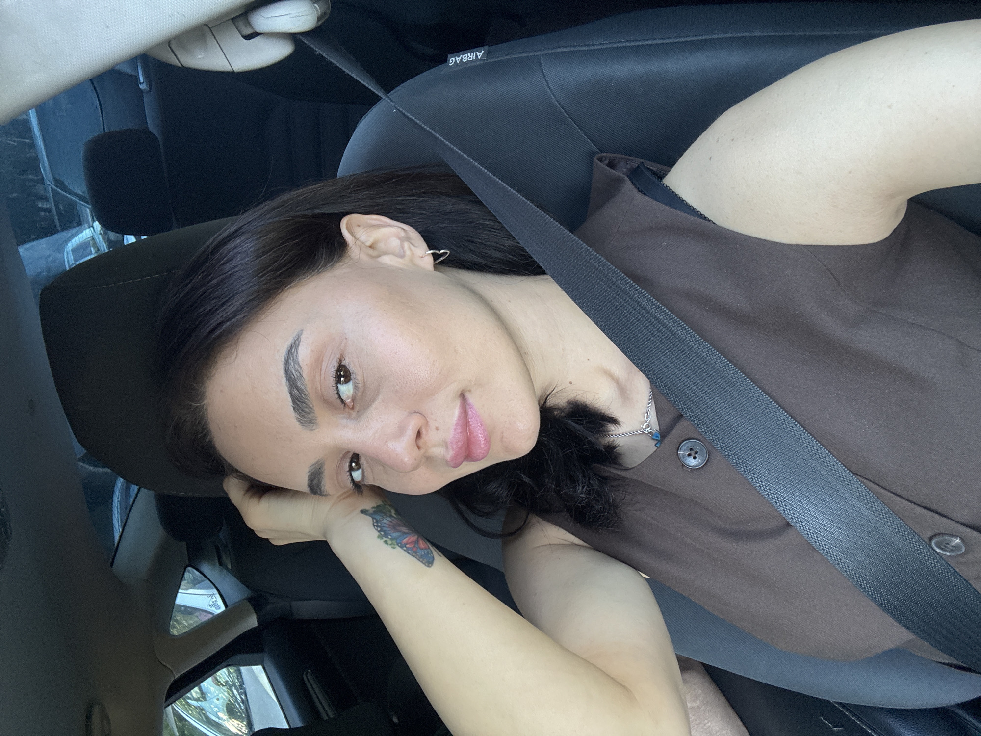A woman with dark hair and brown eyes, wearing a brown sleeveless top and a silver necklace, is sitting in the driver's seat of a car. She has a small butterfly tattoo on her left forearm and a heart-shaped earring in her right ear. A seatbelt is fastened across her chest. The interior of the car is dark, with black seats and a dark headliner. The exterior of the car, visible through the windows, shows trees and other vehicles. The scene appears to be a daytime photograph taken from inside the car. The image does not provide specific details that would allow for identification of the location within Tbilisi, Georgia.
