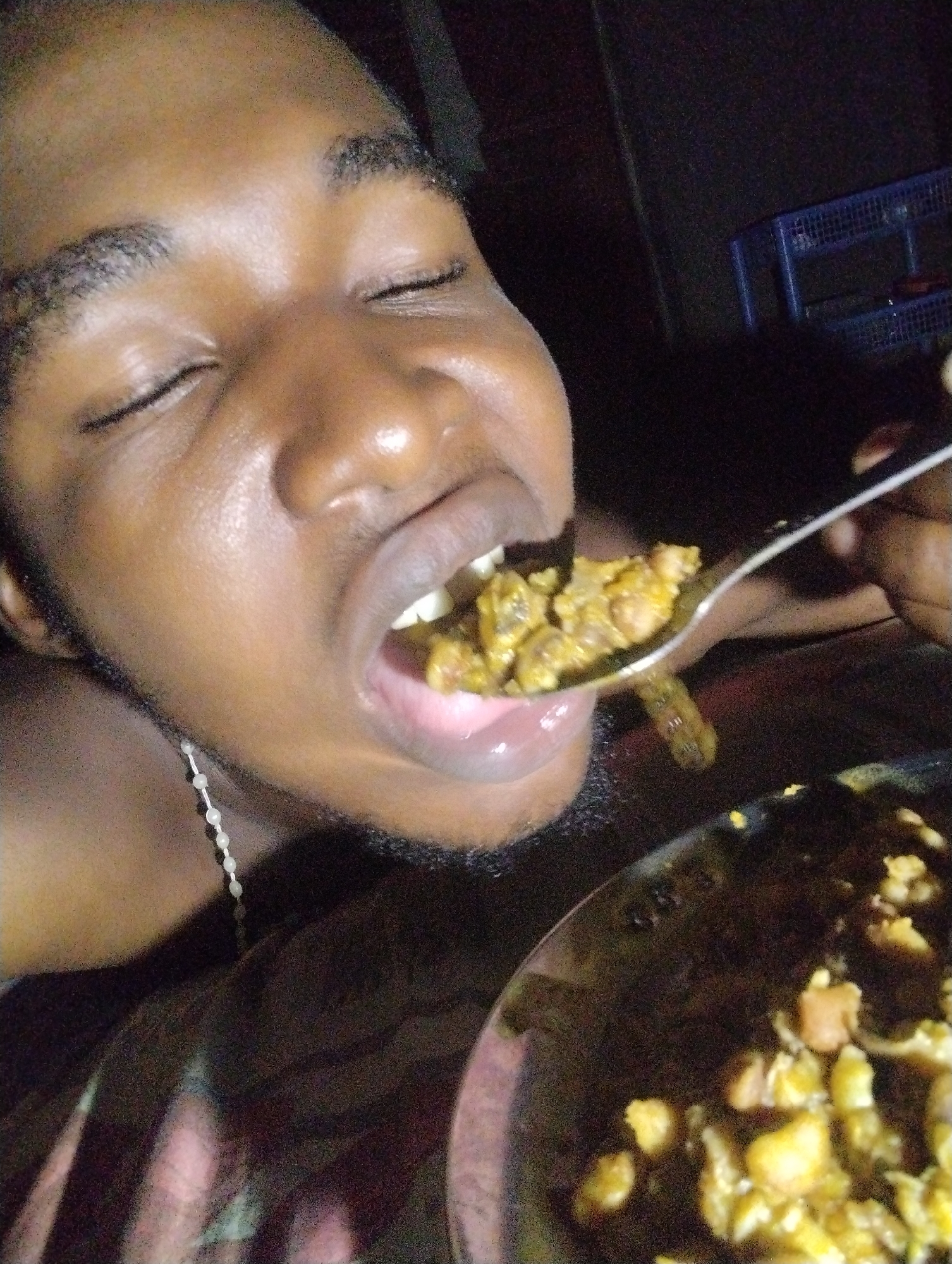 A close-up, low-angle shot shows a person with their eyes closed, about to eat from a spoon. The person's mouth is open, and the spoon, held by their right hand, contains a portion of food. The food appears to be a mixture of small, yellowish-brown pieces, possibly beans or grains in a sauce. A dark plate with more of the same food is visible in the lower right corner. The person is wearing a thin, white beaded necklace. The background is dark and blurred, with a faint glimpse of a blue, slatted shelf unit in the upper right quadrant, suggesting an indoor environment, potentially in Ihiagwa, Nigeria. The overall lighting is dim, with highlights on the person's skin.