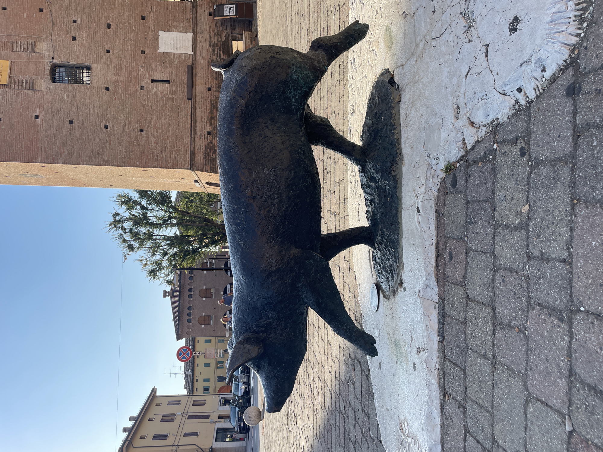 In the city of Castelnuovo Rangone, Italy, a bronze sculpture of a pig is positioned on a textured stone base in a public square. The sculpture depicts the pig in mid-stride, with its front left leg extended forward and its hind legs trailing behind. The pig is dark in color, with a rough texture that suggests age and weathering. The stone base is a mix of white and gray, with visible cracks and an uneven surface. The square itself is paved with gray cobblestones, and a large, reddish-brown brick tower stands prominently in the background. The tower has a square window with iron bars and several smaller openings scattered across its facade. Adjacent to the tower are buildings with yellow and pale orange facades, featuring arched windows and tiled roofs. The sky is clear and blue, with a bright sun casting shadows. Several cars are parked along the street in the background, and two individuals, a man and a woman, are walking together in the middle ground. A traffic sign with a red circle and a diagonal line is visible on a pole in the distance. The overall scene suggests a sunny day in a historic town square.