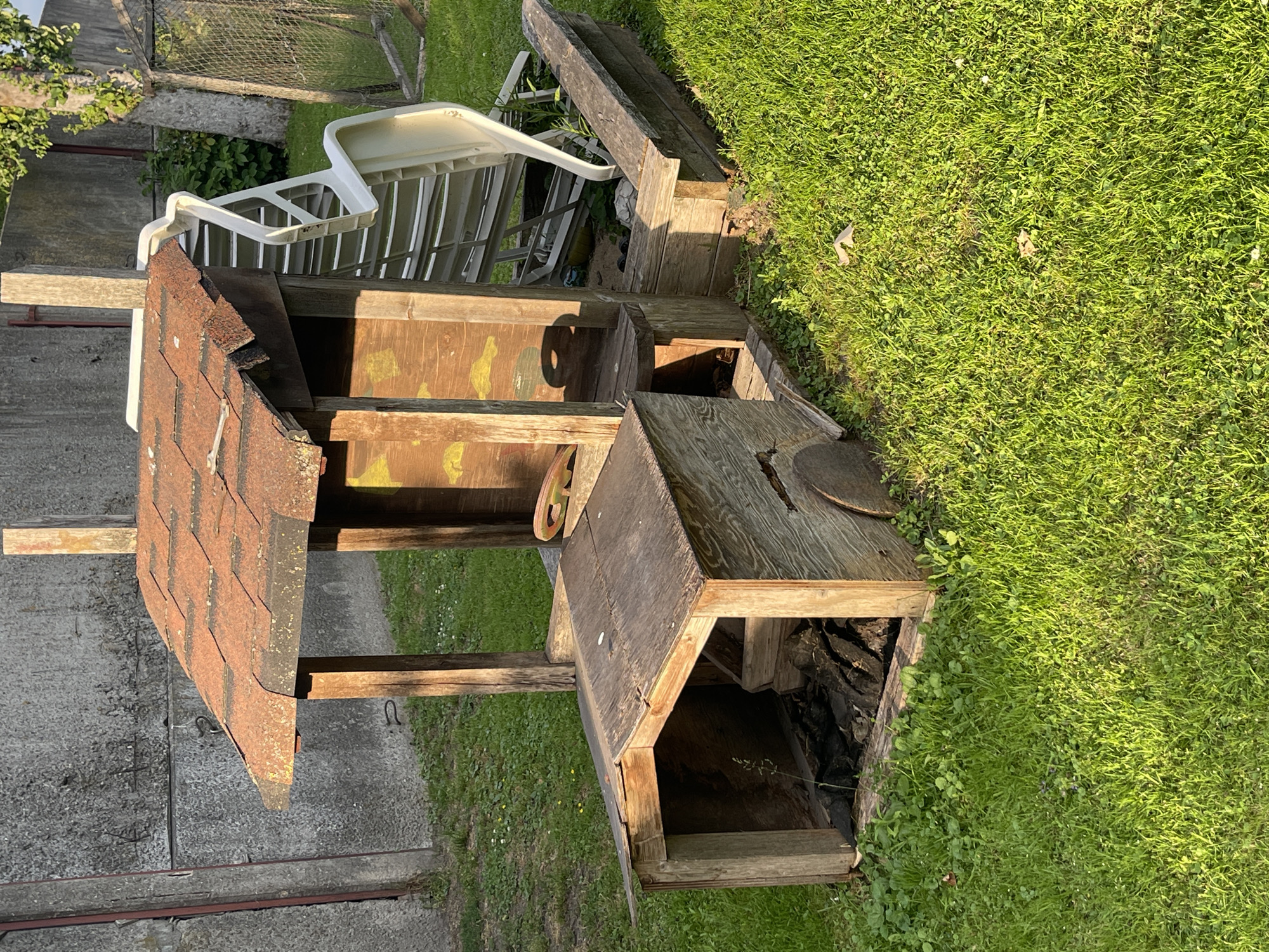 The image shows an outdoor scene in Jelgava, Latvia, bathed in the warm light of what appears to be late afternoon or early evening. The primary subject is a weathered wooden structure, possibly a playhouse or an abandoned shed, situated on a grassy area. This structure has a pitched roof covered with what looks like rusted asphalt shingles. It features open sections, revealing the interior which appears to be empty or filled with debris. A small, rusty metal wheel is visible attached to the side of the structure.
Adjacent to the wooden structure, a white plastic chair is placed on top of other wooden elements, possibly part of a disassembled structure or debris. The background consists of more grass, a concrete wall or fence in the lower left, and a chain-link fence with vegetation behind it. The overall impression is one of disuse and perhaps neglect, with the natural elements of grass and greenery encroaching on the man-made objects. There is no visible text in the image. The lighting suggests a calm, possibly summer evening.