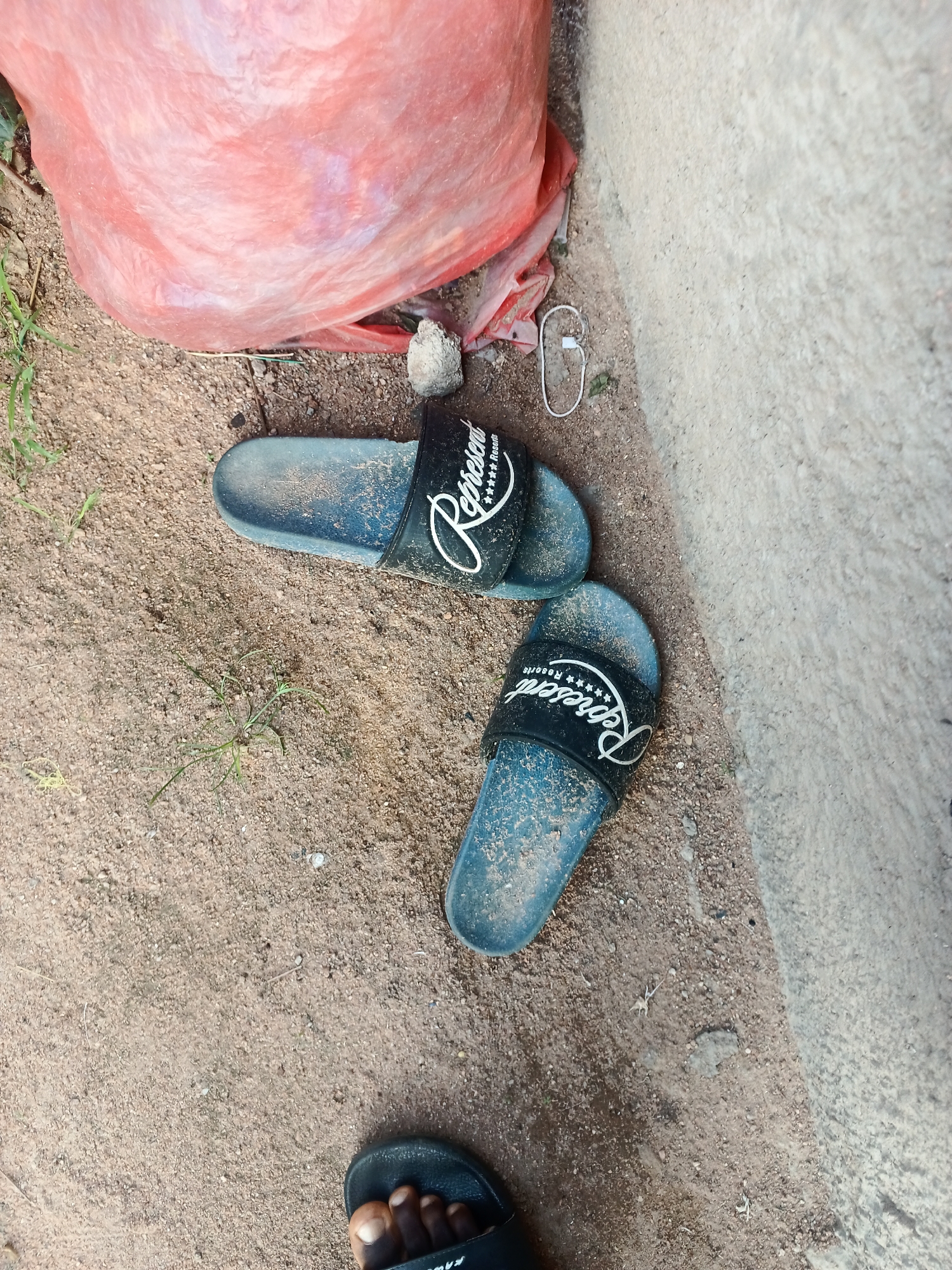 A top-down shot shows a pair of blue slide sandals with black straps scattered on a sandy ground. The straps have the word "Represent" in white lettering with star graphics above and below it. There is a red plastic bag in the upper left corner. A light-colored concrete wall is on the right side of the image. The ground is mostly sand with some small pebbles and sparse green vegetation. A person's foot, wearing a black sandal, is visible in the lower part of the frame. The scene appears to be outdoors, possibly in a dry or dusty environment, with natural lighting. There are no specific cues to indicate the time of day or weather conditions.