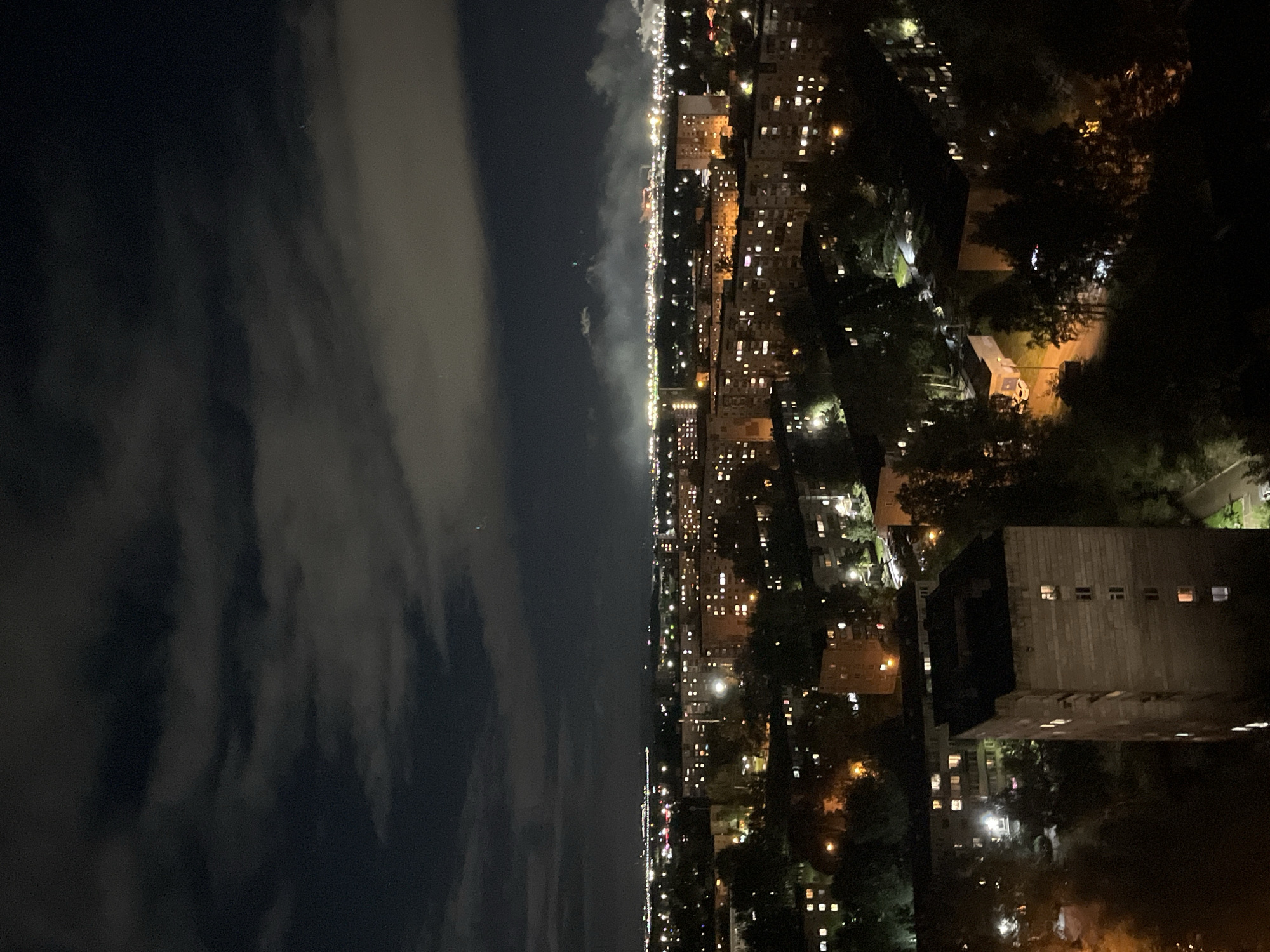 The image is an aerial night view of a city, likely Perm, Russia, showing a dense urban landscape with apartment buildings and trees. The sky is dark with scattered clouds that are illuminated from below by city lights. In the distance, there are faint lights suggesting further development or industrial activity, with plumes of smoke or steam rising. Numerous windows in the apartment buildings are lit, indicating residential occupancy. The foreground features a taller, concrete building with visible windows and a dark, possibly flat roof. The overall impression is one of a typical nighttime scene in a residential area of a Russian city.