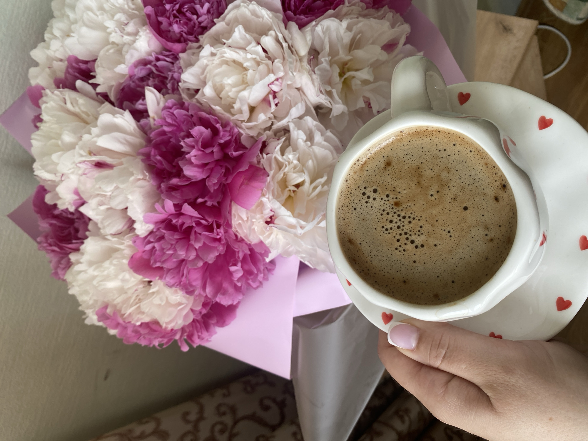 A medium-angle shot depicts a bouquet of peonies and a cup of coffee held by a person's hand. The bouquet consists of both white and pink peonies, wrapped in light pink paper. The cup of coffee is white with small red hearts adorning its surface, and is filled with a dark brown beverage with foam. The hand holding the cup has pale skin and a light pink manicure. The background consists of a light-colored wall and part of a patterned sofa or chair. The location is indicated to be Sochi, Russia, but there is no visual indication of this location in the image.