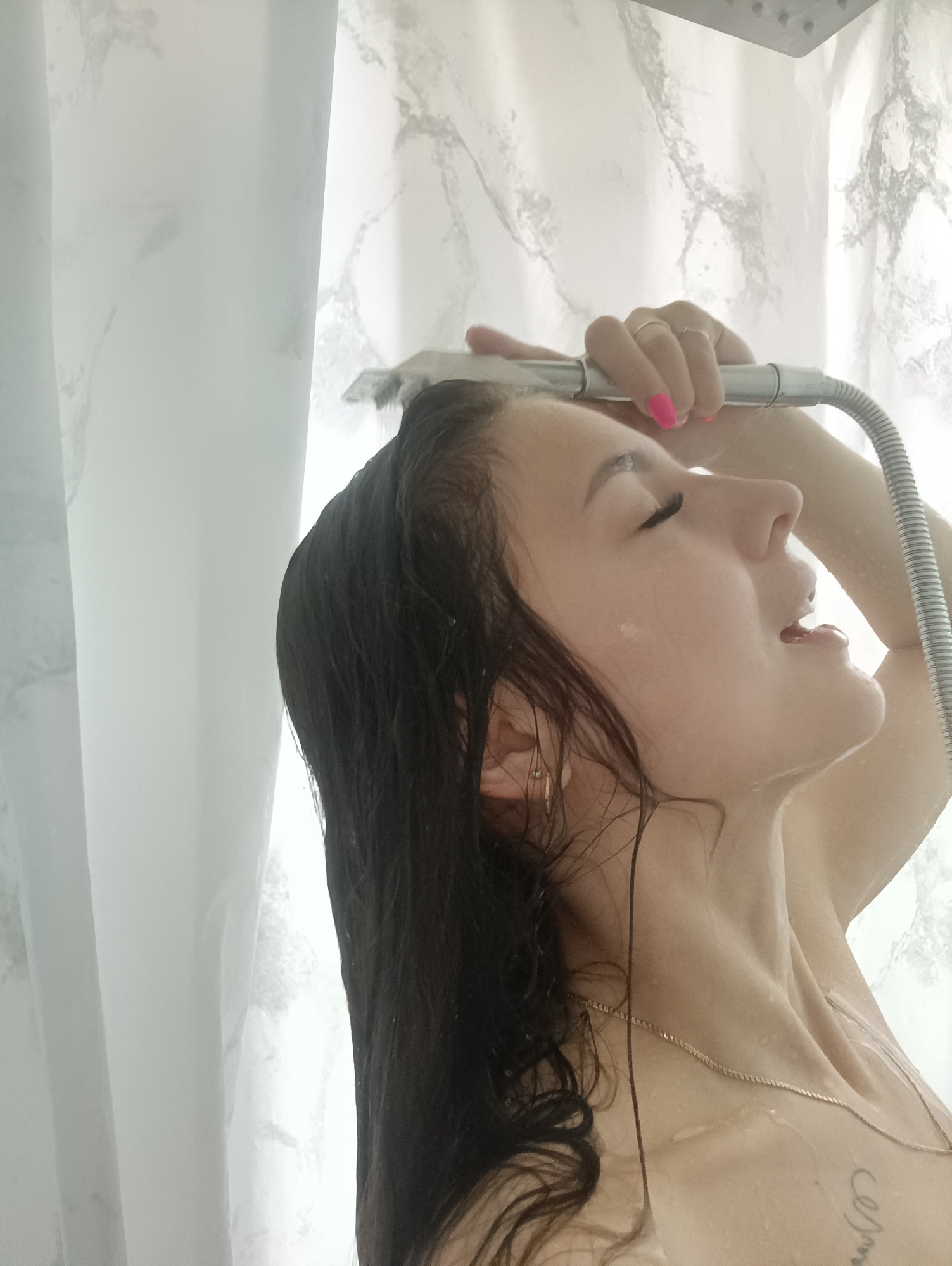 A person is in a shower with a white, marble-patterned shower curtain. They are holding a handheld shower head above their head, and water is running over their face and hair. The person has dark, wet hair, fair skin, and their eyes are closed. They are wearing a thin gold necklace. A small gold hoop earring is visible in their left ear. The person's nails are painted pink. A tattoo is visible on their chest.