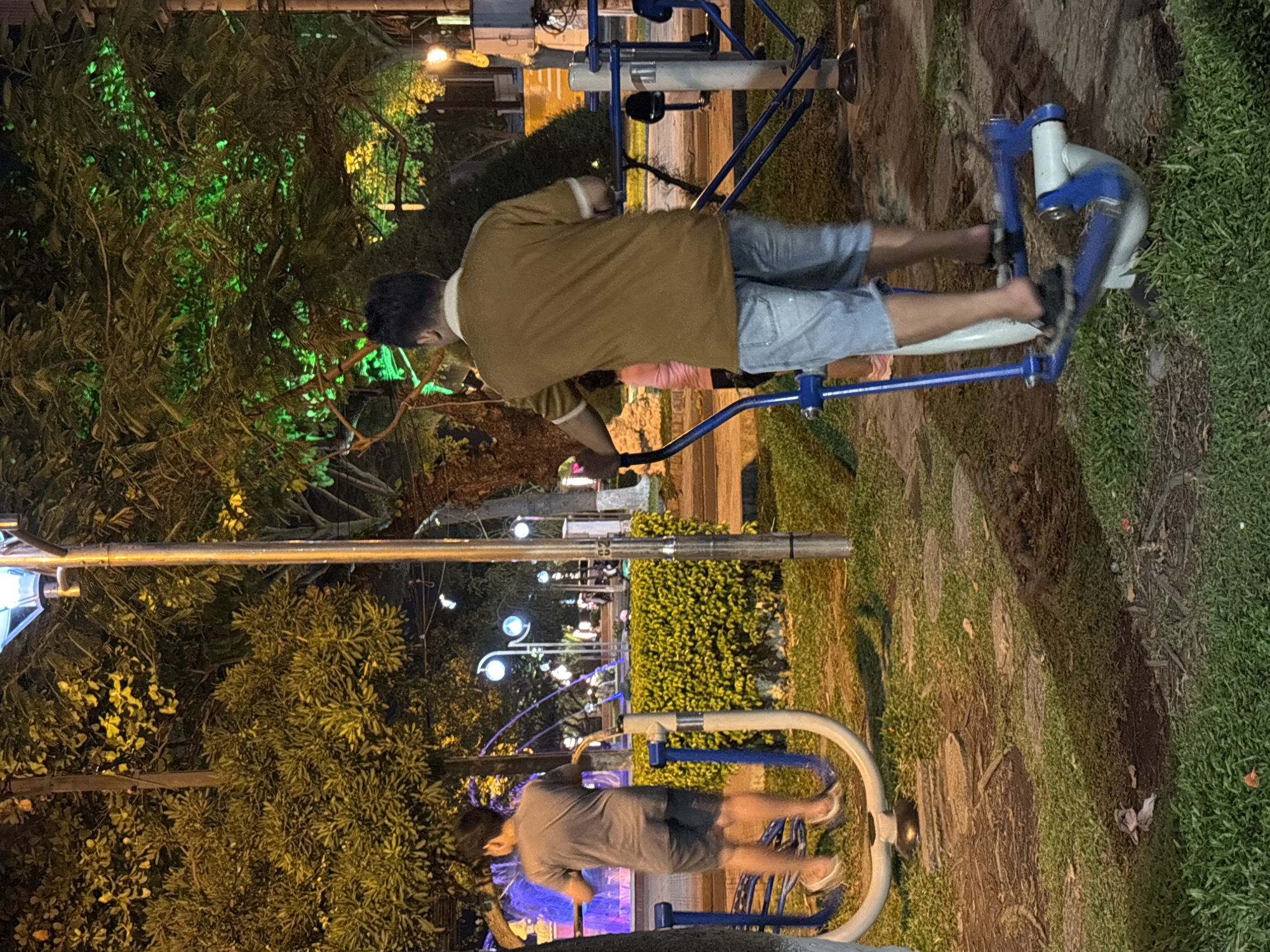 The image depicts an outdoor fitness park at night in Nha Trang, Vietnam. In the foreground, a man wearing a brown polo shirt and denim shorts is actively using an elliptical machine. To his left, a younger person in a grey t-shirt and shorts is on a similar elliptical machine, facing away from the camera. The park features green trees, some illuminated with green lights, and a fountain with blue and purple lights in the background. Several lampposts are visible, providing light to the scene. The ground is a mix of grass and stone pathways.
