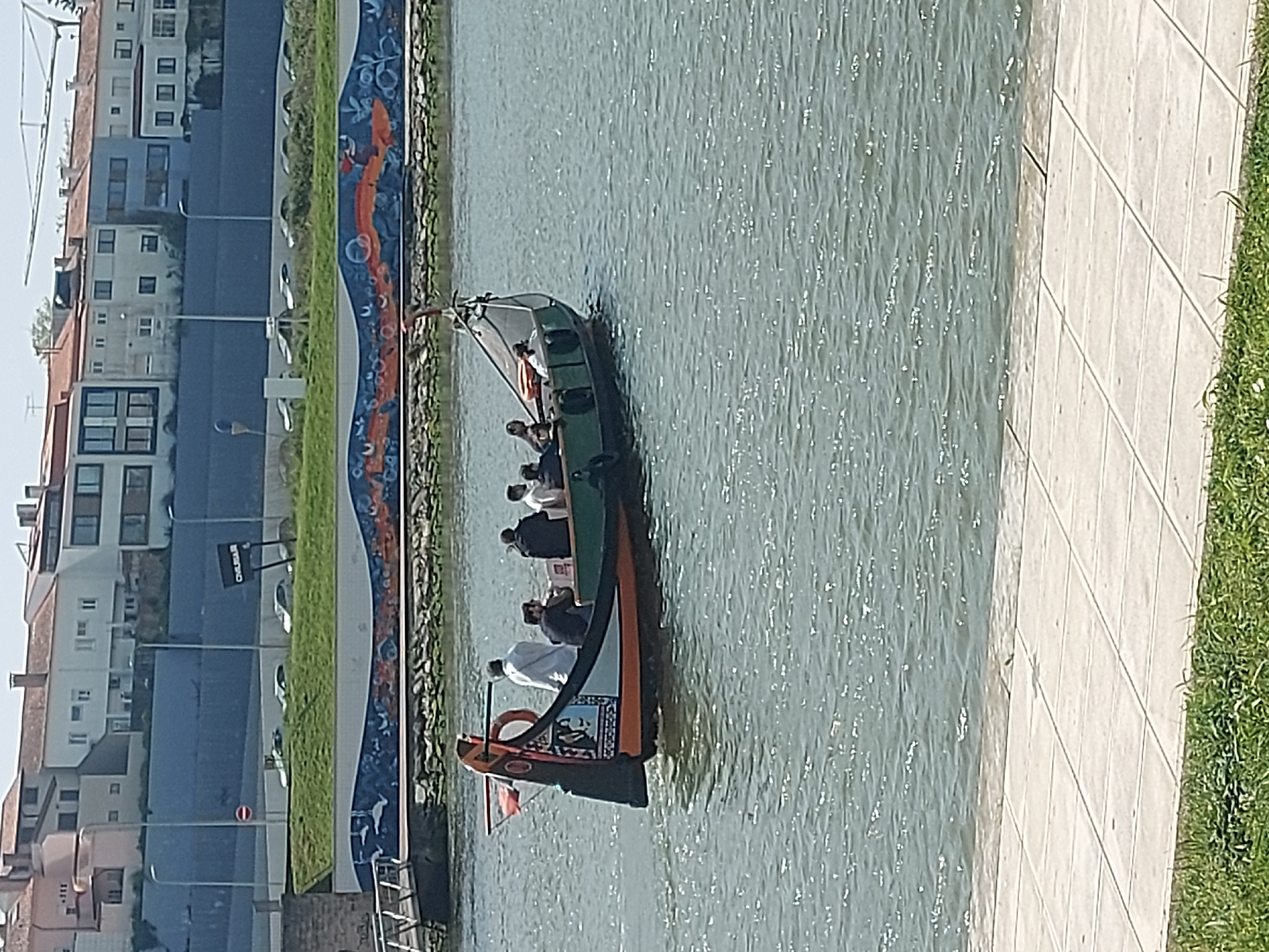 The image depicts a moliceiro boat navigating a canal in Aveiro, Portugal. The boat is carrying multiple passengers. Adjacent to the canal is a pedestrian walkway composed of tiled pavers and a strip of grass. Buildings are seen in the background on the other side of the canal. An artistic mural decorates the canal wall.