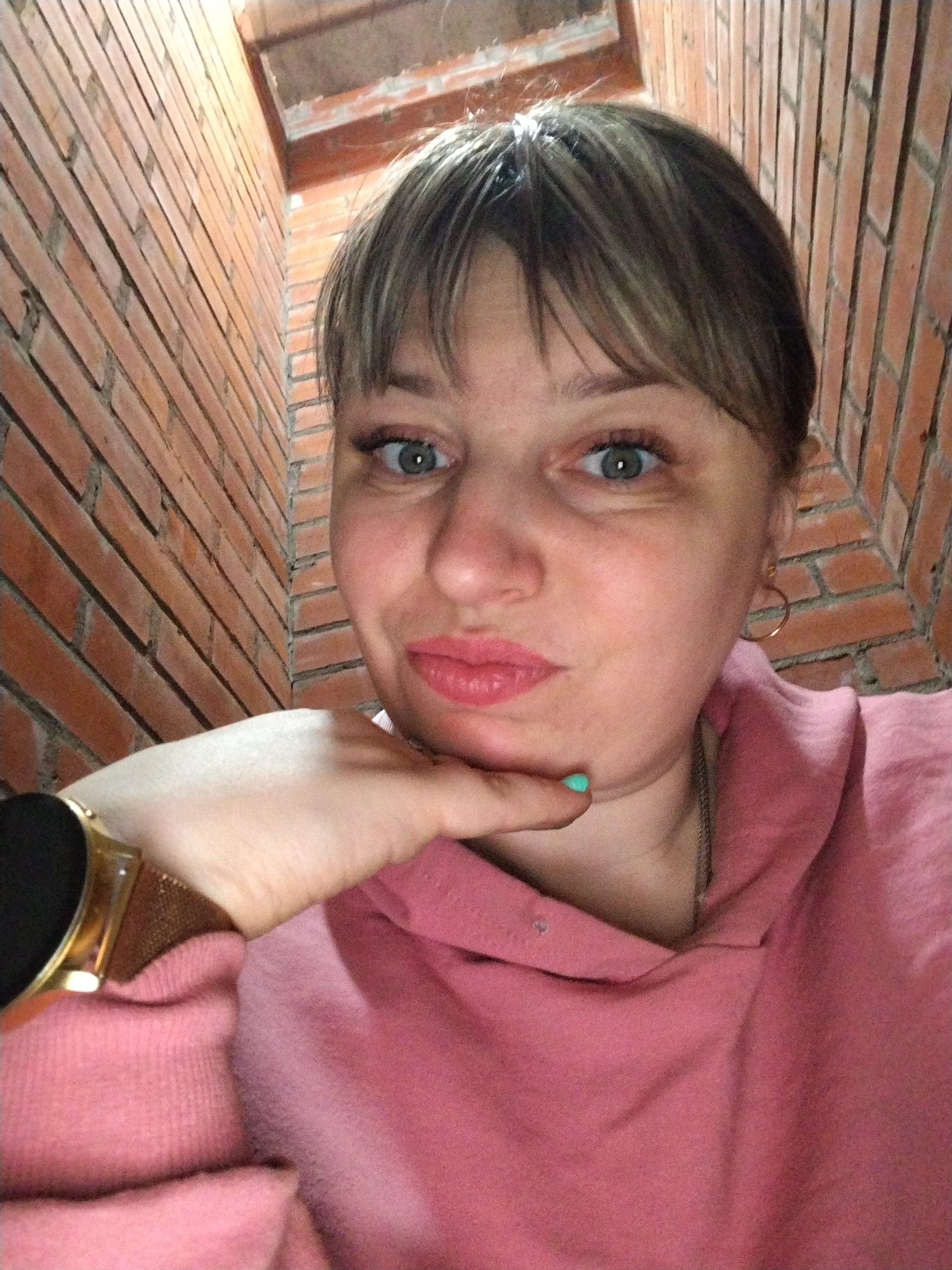 A close-up selfie depicts a woman looking directly into the camera. She has light-colored hair with bangs, light green-blue eyes, and is wearing pink lipstick. Her chin is resting on her left hand, which has a bright turquoise fingernail polished on the index finger. She wears a pink hooded sweatshirt (hoodie). A gold-colored watch with a textured strap is visible on her left wrist, and a thin gold hoop earring is in her right ear, along with a delicate silver-toned necklace.
The background features a prominent red brick wall, which appears to be an interior surface. The angled perspective suggests a corner or a narrow space, possibly a hallway or stairwell within a building. The lighting is bright, illuminating both the woman's face and the brickwork.
The woman displays a slightly amused or playful expression, with her lips gently parted and her eyes wide. The image captures an intimate moment, likely taken indoors in Шебунино, Russia. No visible text is present.
