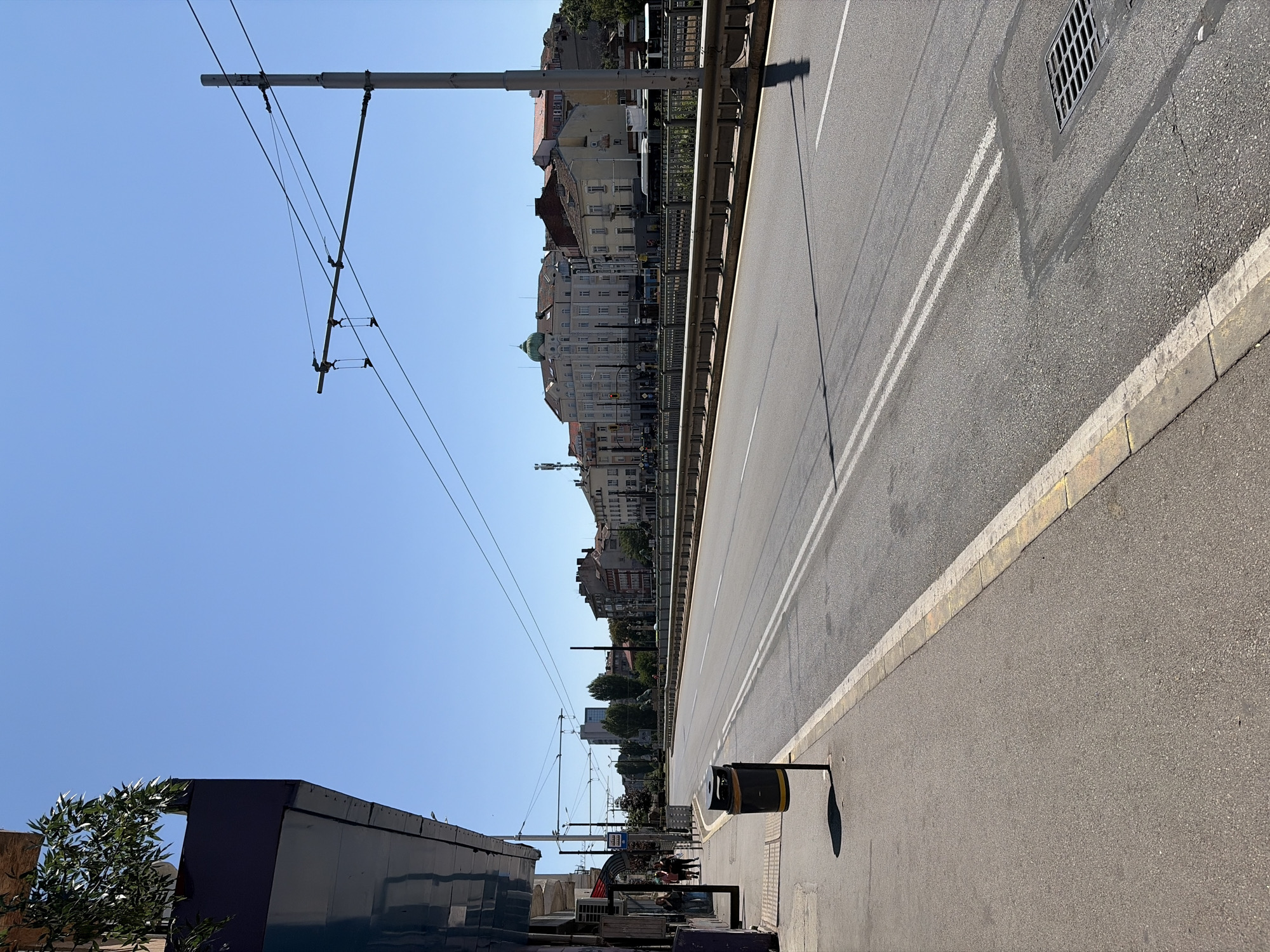 The image shows a wide, paved street in a city, likely Sofia, Bulgaria, under a clear blue sky. On the left side of the street is a sidewalk with a building facade featuring reflective glass and a balcony with greenery. A trash bin stands on the sidewalk near the building. Several people are walking on the sidewalk. The street has double white lines marking the lanes and appears to be a bridge or elevated roadway, as a metal railing runs along its right side, separating it from a lower area or riverbed not fully visible. In the distance, across the street, is a row of multi-story buildings with varied architecture, including some with prominent domes and balconies. Overhead, there are electrical wires and poles, suggesting public transportation like trams or trolleybuses. A drainage grate is visible on the road surface on the right side of the image. The scene is brightly lit by sunlight, casting shadows from the objects and structures.