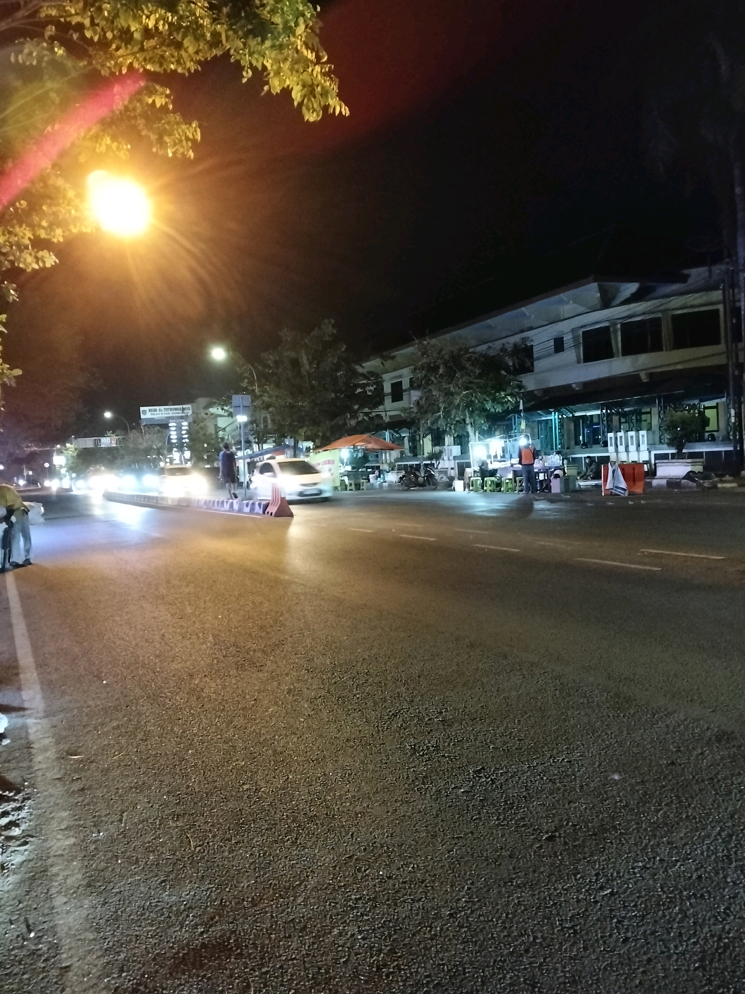 This is a nighttime photograph of a street scene in Purworejo, Indonesia. The image captures a busy intersection illuminated by streetlights and the headlights of passing vehicles. In the foreground, the asphalt road is visible with white lane markings. On the right side of the road, a building with multiple windows and a covered awning is situated. Several street vendors are operating in front of the building, with food stalls and seating areas set up. A few individuals are seen interacting with the vendors or walking along the sidewalk. A white car is stopped in the middle of the intersection, and other vehicles are driving by, their headlights creating streaks of light. In the background, trees and foliage can be seen, along with more streetlights. The overall atmosphere is one of urban activity at night.