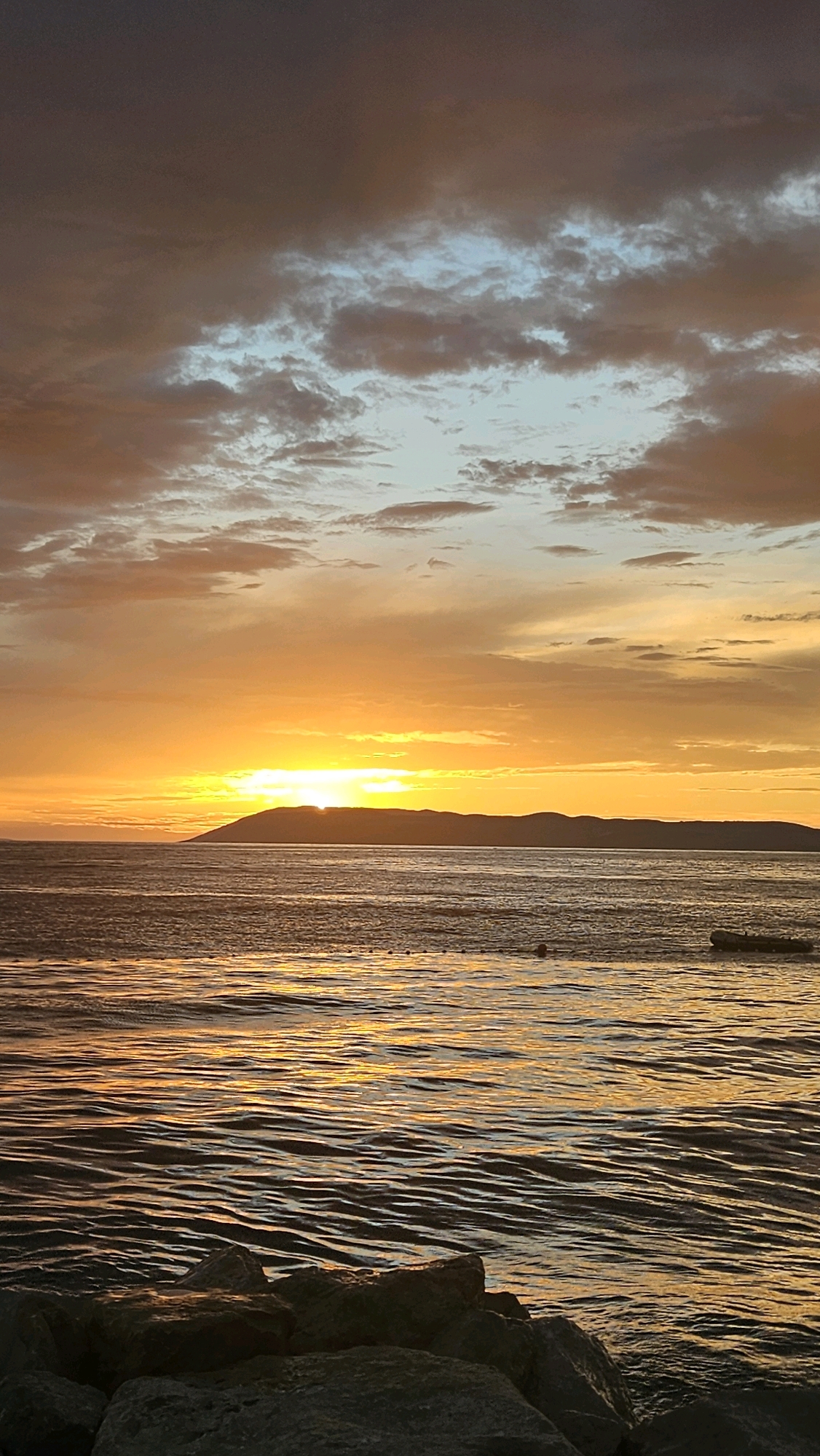 A vertical, eye-level, full shot captures a sunset over the sea at Tučepi, Croatia. The sun is low on the horizon, casting a bright orange glow across the sky and reflecting on the rippling water. A dark, low-lying island is silhouetted against the vibrant sky. The water in the foreground is choppy, with golden reflections. The bottom of the frame is occupied by large, dark rocks, presumably part of a breakwater or shoreline. The sky is filled with dramatic clouds, ranging from dark gray to light blue and orange, illuminated by the setting sun.