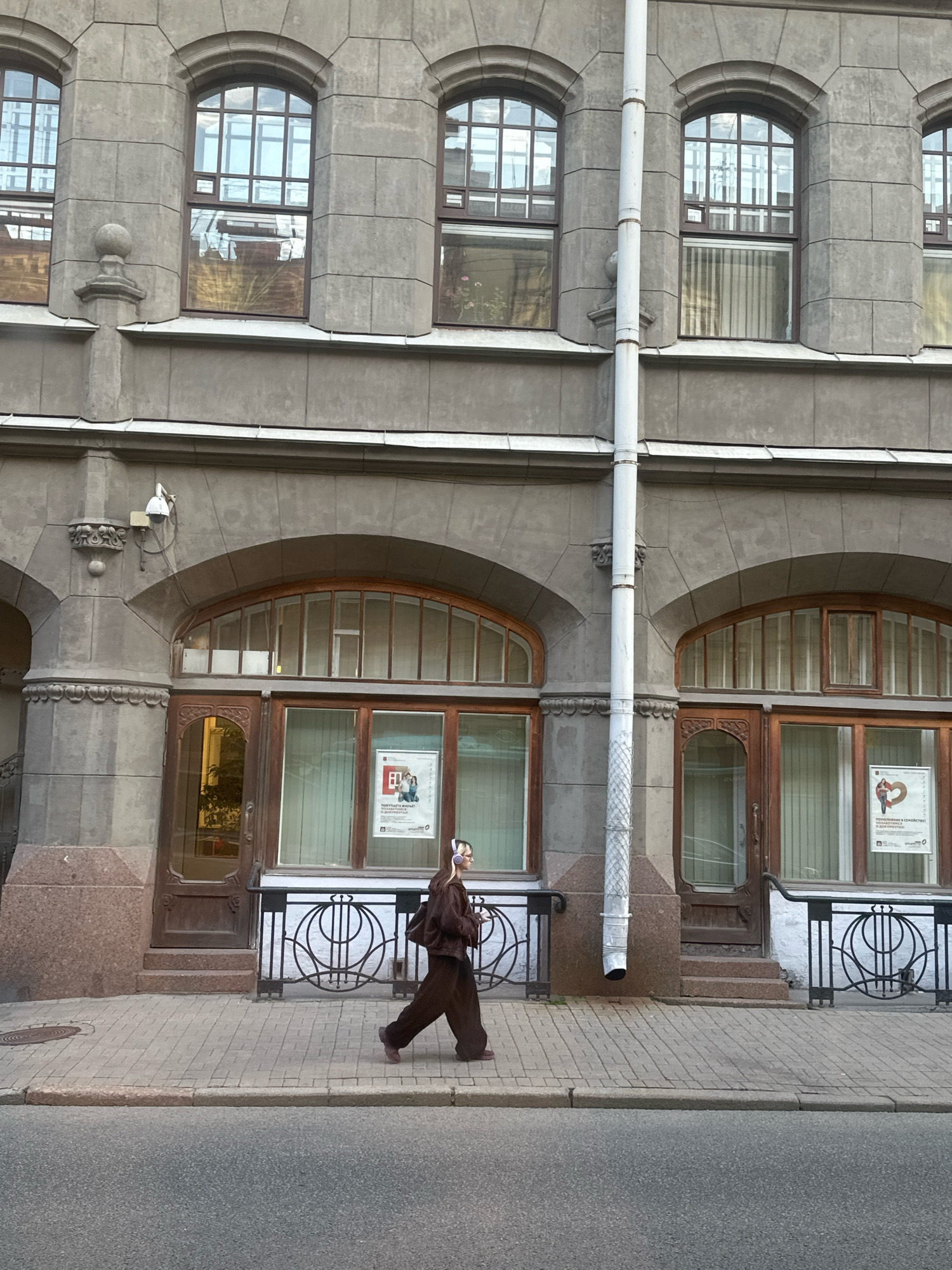 A person walks along a paved sidewalk in front of a building with arched windows and doors. The building appears to be made of stone and has a white pipe running down its facade. The person is wearing dark clothing and headphones. There are posters visible in the windows. The scene is lit by natural daylight. Based on the architectural style, the location is likely Saint Petersburg, Russia.