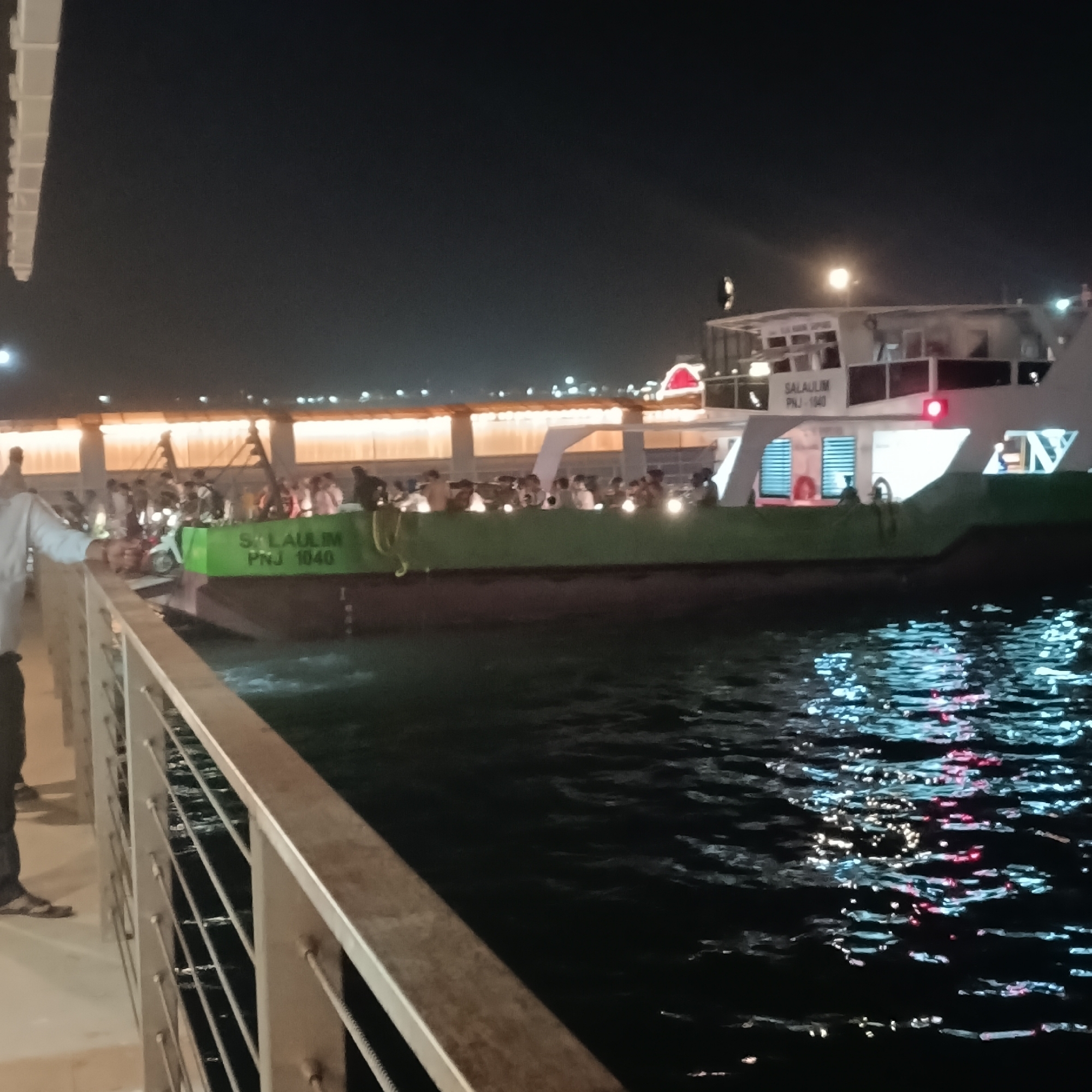 At night, a multi-deck ferry boat, "SALAVULIM PNJ-1040", is docked at a waterfront in Panaji, India. Passengers stand on the main deck. A person stands next to a metal railing in the foreground. The water reflects light. A building with lights lines the waterfront.