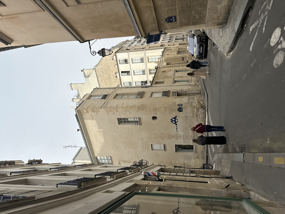 The image depicts a narrow street intersection in Paris, France, specifically Rue Guénégaud, under an overcast sky. Historic, multi-story buildings with light-toned stone and stucco facades line both sides of the street. On the left, a building features a storefront displaying a red, white, and blue barber pole, with a bicycle parked near its entrance. The prominent central building, light-tan in color, exhibits two blue pixelated "Space Invader" artworks on its facade; one larger, and a smaller one to its left.
Four individuals are present on the street, all walking away from the viewer. In the mid-ground, two women walk side-by-side; one wears a long black dress, and the other a red top with blue jeans. Further down the street on the right, two additional individuals, one in a light trench coat and another in a dark jacket, are also walking. A black Audi sedan is parked on the right side of the street, adjacent to a building with an ornate black street lamp affixed to its facade. A blue street sign on this building reads "18 Rue Guénégaud". The asphalt street surface shows partial white crosswalk markings and a faded white bicycle lane symbol.