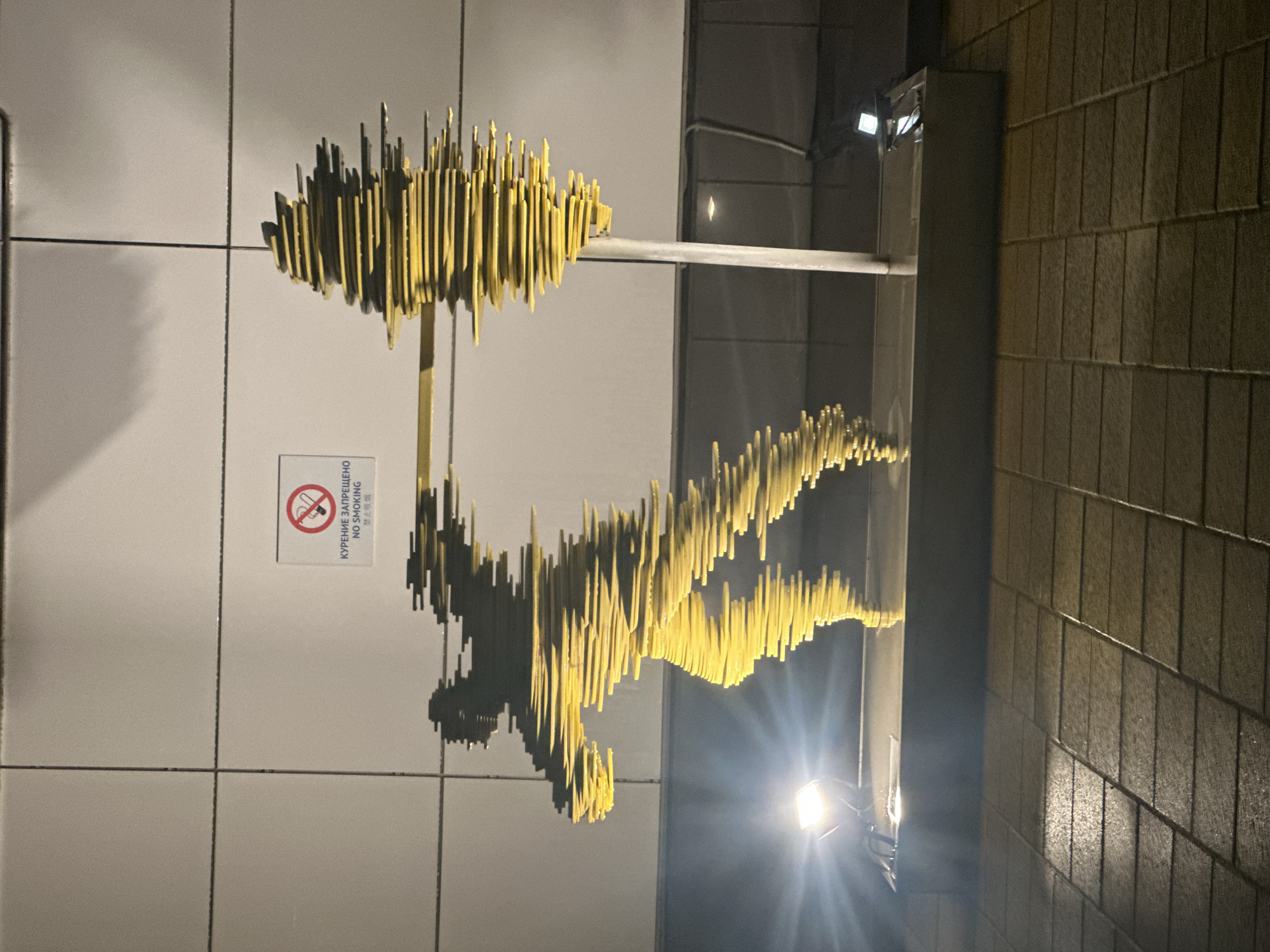 A sculpture is mounted on a building, located near a "no smoking" sign written in both Russian and English. The sculpture, primarily yellow in color, appears to be constructed from multiple vertical elements arranged to suggest a human figure and a separate cloud-like form connected by a horizontal beam. The sculpture is illuminated by nearby lights. The building features a mix of tiled and brick exterior surfaces. The location is Stavropol, Russia.