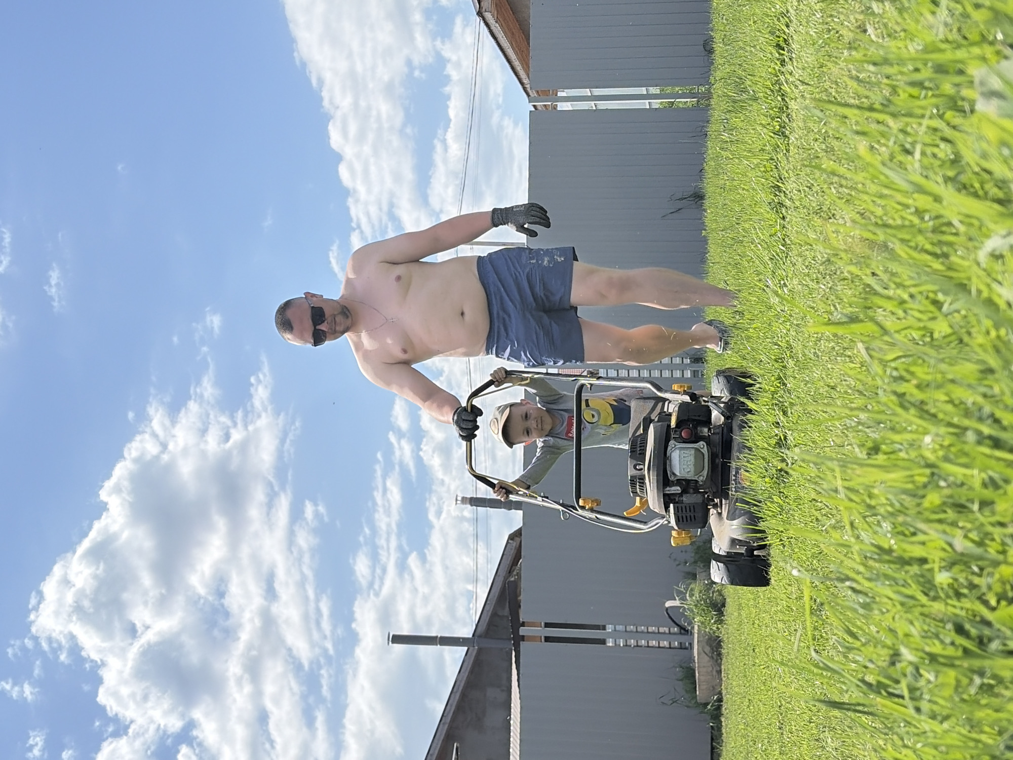 A shirtless man wearing sunglasses and shorts is mowing a lawn with a gas-powered lawnmower. A child in a gray T-shirt is standing in front of him, holding the lawnmower's handle. The man is wearing one black glove. The lawn is green and tall, and a gray fence and building are in the background. The sky is blue with scattered white clouds. The photograph may have been taken in Chaikovsky, Russia, but there is no definitive evidence of this in the image.
