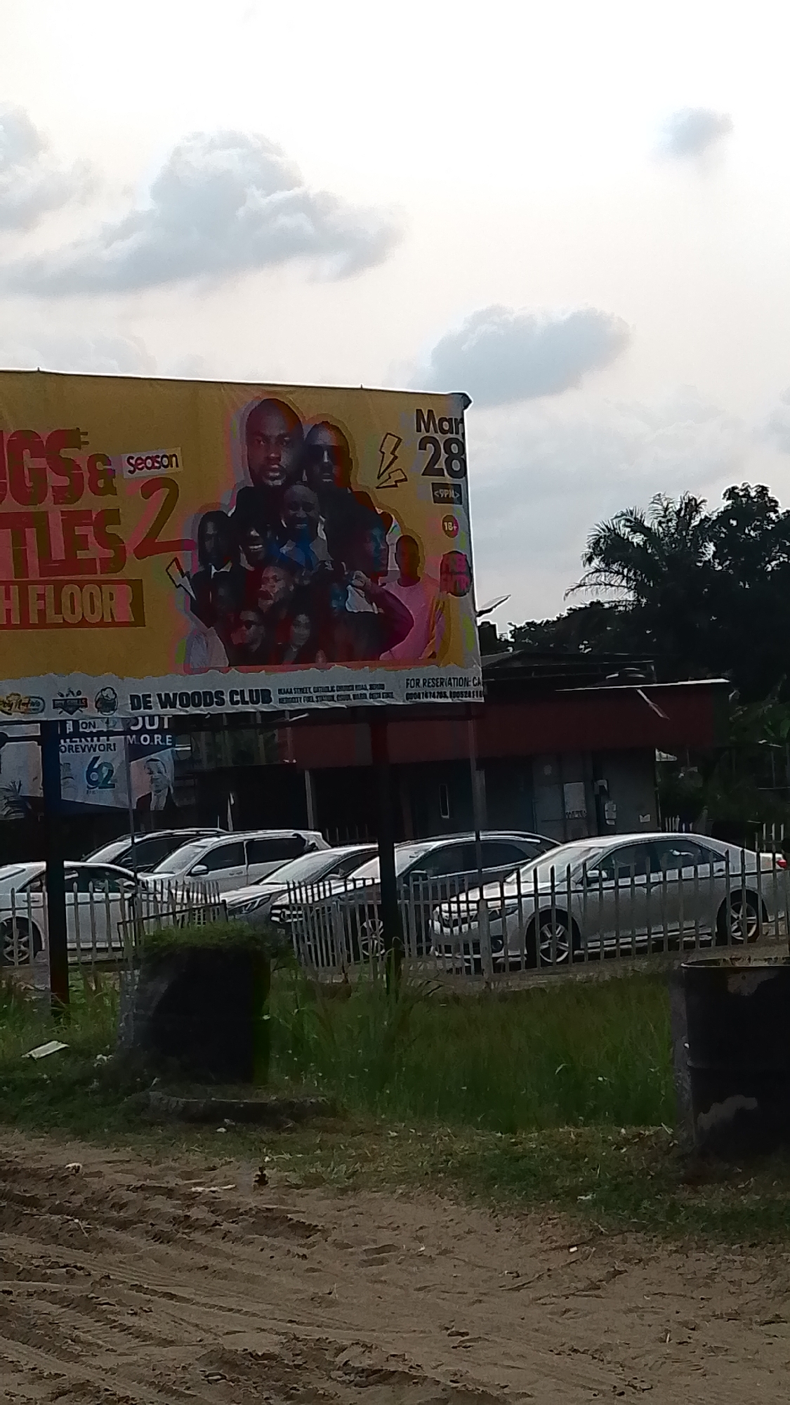 The image shows an outdoor scene in Warri, Nigeria. A large billboard displays promotional content for a "Season 2" event at "De Woods Club," scheduled for March 28 at 9 PM, with an 18+ age restriction. The club's address is listed as "Kaara Street, Catholic Church Road, Warri," with "Mercy City Fuel Station" as a nearby landmark. Reservation contact numbers are provided. The billboard also features images of several individuals.
Below the billboard is a parking area with several silver-colored vehicles behind a metal fence. Vegetation grows between the fence and a dirt road in the foreground. There is an object along the road. The sky is cloudy.