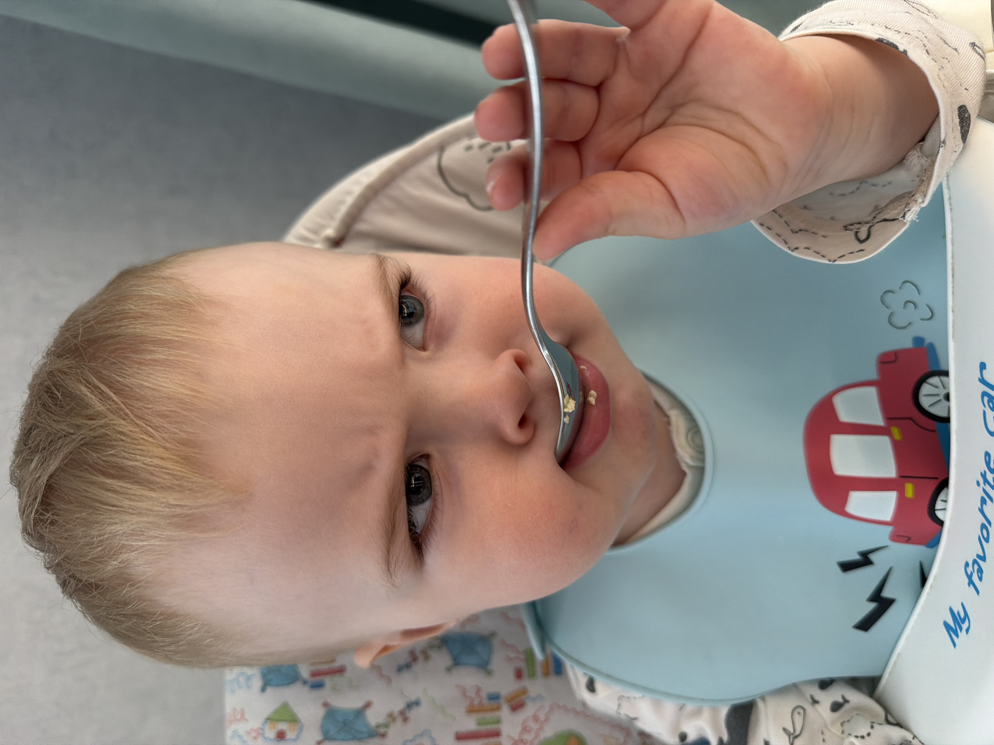 A baby wearing a light blue bib with a red car design and the text "My favorite car" is being fed with a silver spoon. The baby is lying down, with their head turned to face the camera. The background appears to be a light-colored surface with patterns. The location is unknown and cannot be determined from the image.