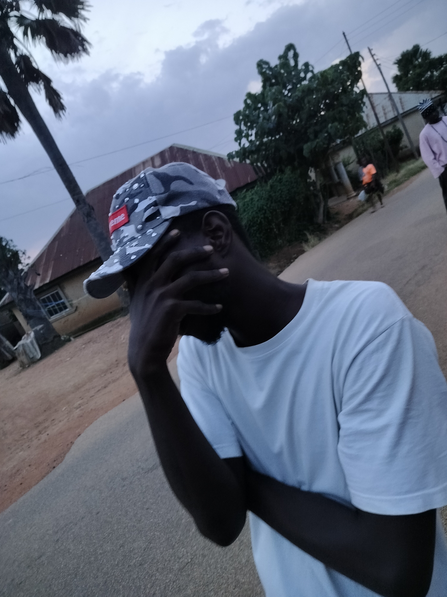 The image shows a young man wearing a white t-shirt and a camouflage-patterned baseball cap with a red square logo. He is standing outside, with his hand covering his face, possibly indicating distress or shyness. In the background, there are buildings with corrugated metal roofs, lush green trees, and a palm tree. A road is visible in the foreground. The sky is overcast. The presence of palm trees and the architectural style of the buildings suggest a tropical or sub-tropical location. The overall impression is of a street scene in a residential area, possibly during dusk given the lighting conditions. The location is identified as Du, Nigeria.