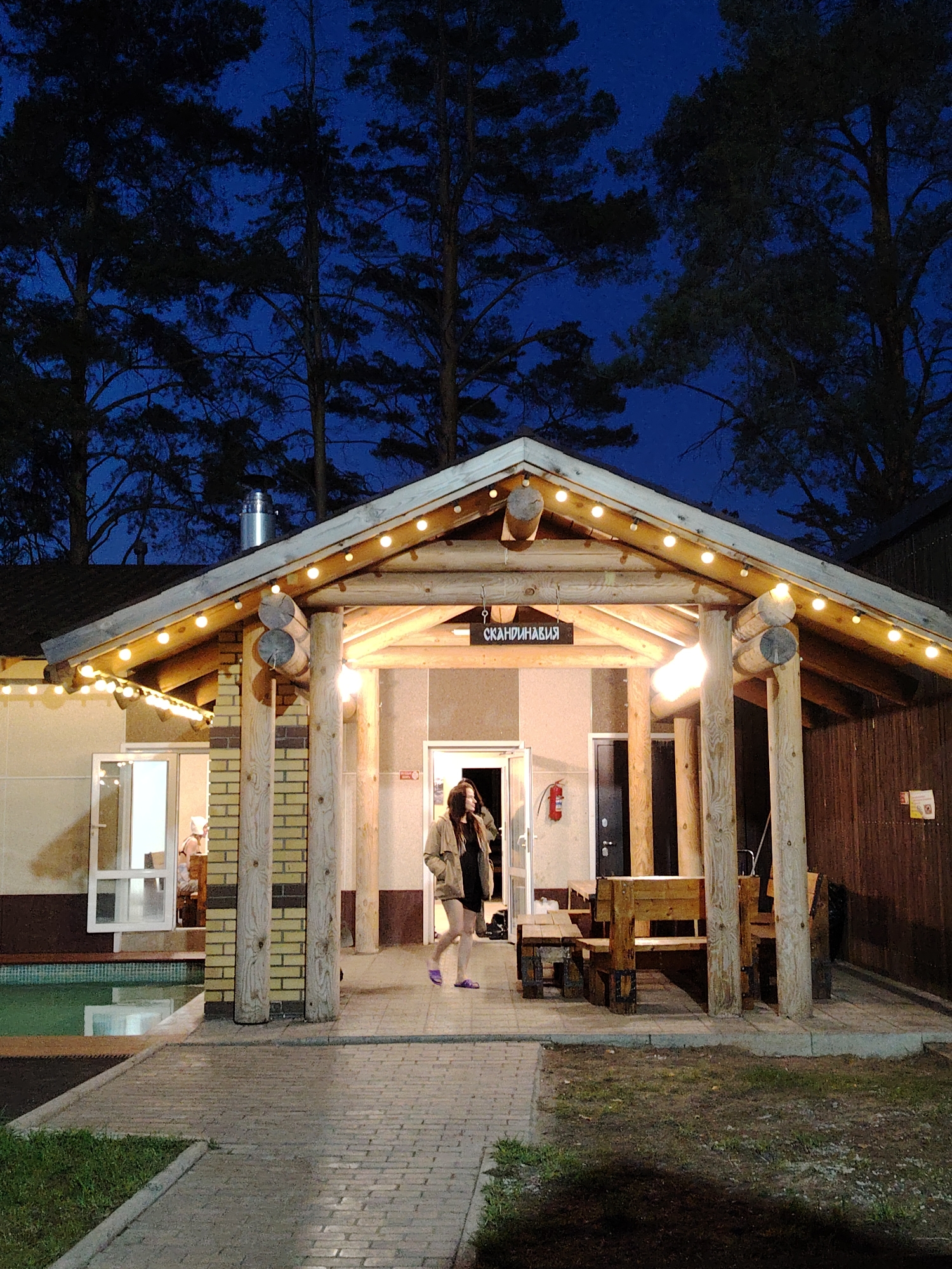 Captured at night in Chaikovsky, Russia, the image presents a single female figure exiting a building marked with the Cyrillic word "Скандинавия." The building features a covered porch illuminated by string lights. Its construction incorporates brick and exposed wooden beams. The female, clad in a light-colored jacket over a dark dress and wearing purple slippers, walks away from the entrance. Inside the building to the left, another individual is visible. A wooden picnic table and bench set sit under the porch. Tall trees are visible in the background against a dark blue sky. A brick-paved walkway leads away from the building.