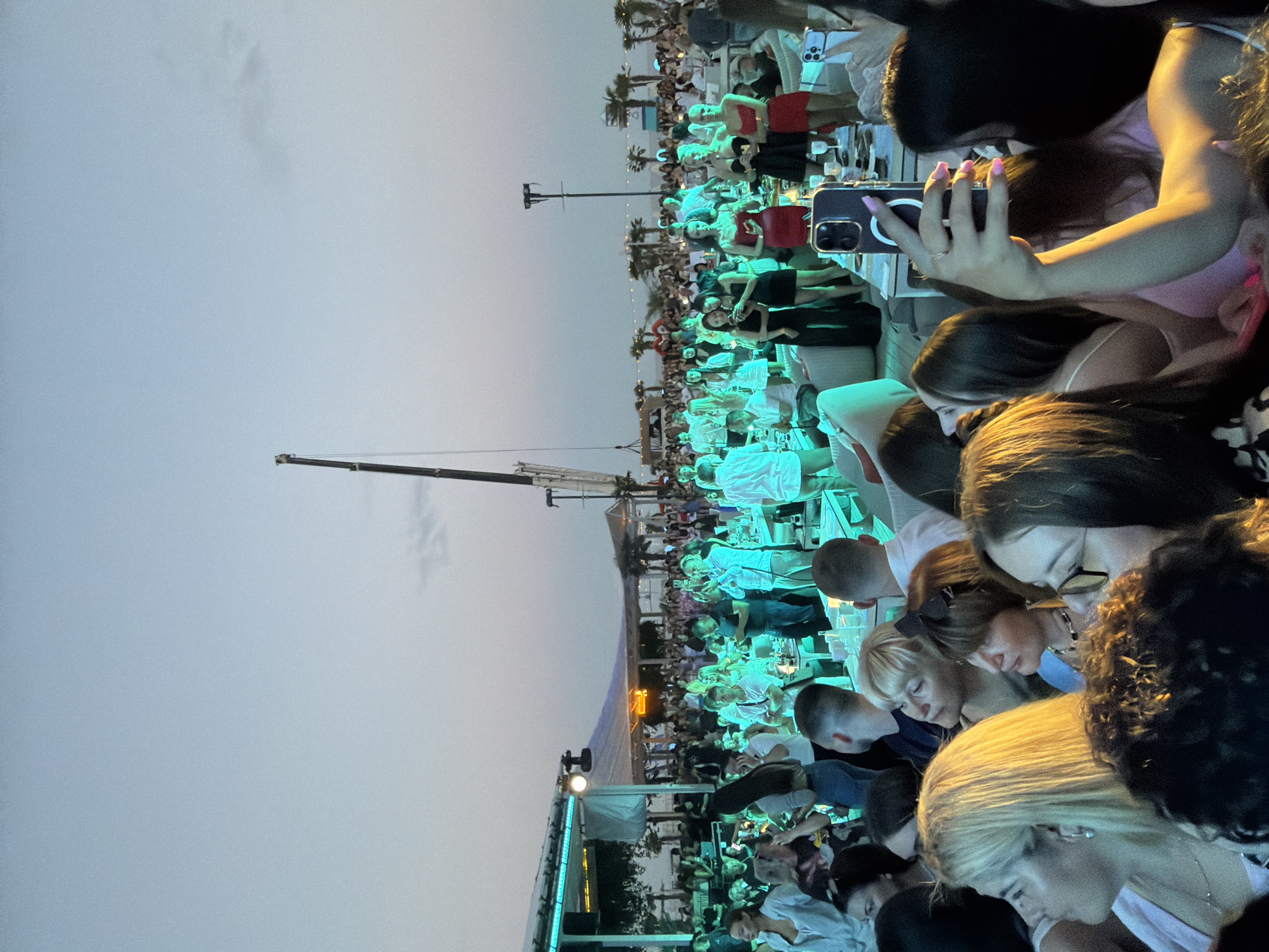An outdoor event is taking place at dusk in Odesa, Ukraine, with a large crowd of people gathered. A construction crane is visible in the background, indicating ongoing development or preparation. Many attendees are holding up their phones, suggesting they are taking pictures or videos of the event. The lighting is predominantly a vibrant green, illuminating a stage area and parts of the crowd. Palm trees are scattered throughout the venue, suggesting a beachside or resort-like setting. Several tables with drinks are visible, indicating a social gathering with food and beverages. Some individuals are dressed in formal attire, including dresses, while others are in more casual clothing. The overall atmosphere appears to be festive and lively, with a sense of celebration.
