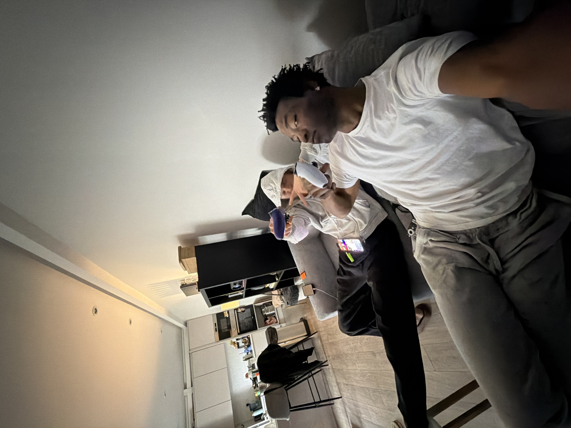 Two individuals are visible in an interior setting, possibly a residential apartment. One individual, a Black male, is closer to the camera, wearing a white t-shirt and grey trousers, lying on a grey sofa. The other, whose face is partially covered, is wearing a white top and black trousers and is holding a purple cup. A black shelving unit against a wall houses several items. A black coat hangs on a chair. The location appears to be a modern apartment, suggestive of an urban area, though specific London landmarks are not visible within the frame to definitively identify the exact location in London.