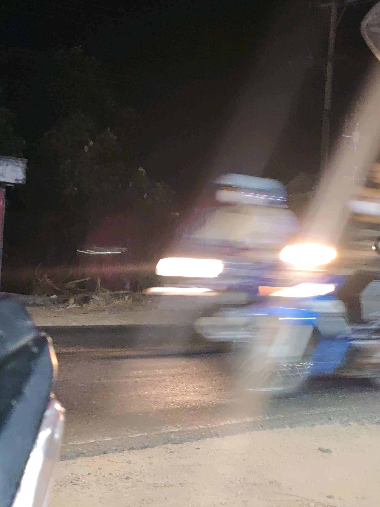 The image shows a blurred, nighttime scene on a road in Gumuk Mas, Indonesia. A motorcycle, moving at speed, is the central subject, its headlights creating bright streaks of light across the frame. The rider is indistinct due to motion blur. The road surface appears to be asphalt, with some gravel and debris visible. To the left, a portion of a dark-colored vehicle, possibly a car, is partially visible, also contributing to the sense of movement. In the background, trees and foliage are suggested by dark masses, and a faint structure, possibly a utility pole, is visible on the right. The overall impression is one of a fleeting moment captured during nocturnal travel.