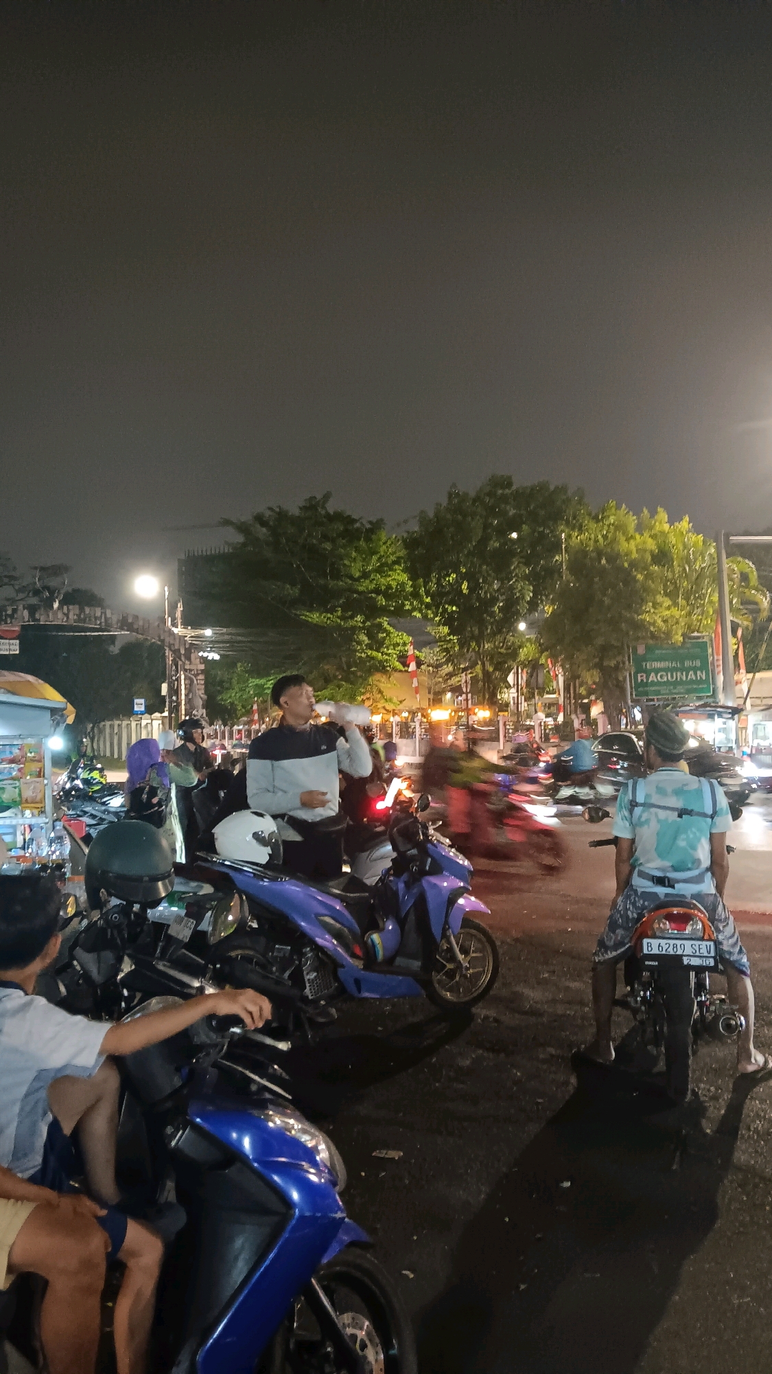 In South Jakarta, Indonesia, at night, several people are gathered around motorcycles on a street. A young man in the center of the frame, wearing a grey and black long-sleeved shirt, is drinking from a clear plastic bottle. He is standing between two parked motorcycles. To his right, another young man wearing a light blue and white patterned shirt is seated on a motorcycle, facing away from the camera. His legs are visible, and he is wearing flip-flops. The background features streetlights, trees, and traffic, indicating an urban environment. A green street sign reads "TERMINAL BUS RAGUNAN". Multiple motorcycles are visible, some parked and others in motion, creating a dynamic scene. The ground is asphalt, and shadows are cast by the lighting.