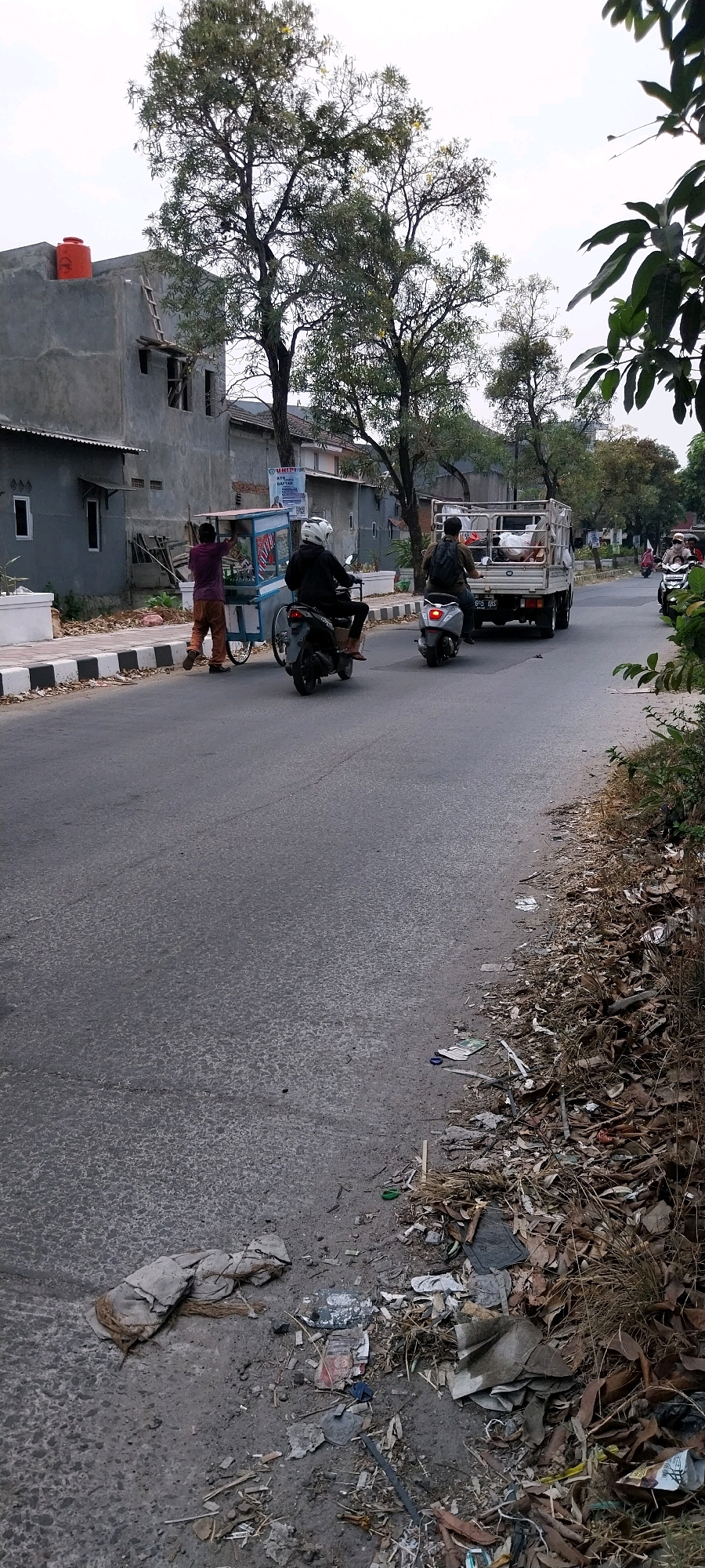 In Bojong Nangka, Indonesia, a street scene unfolds with a gray asphalt road bordered by a sidewalk and a line of trees. On the sidewalk, a man in a purple shirt and brown pants pushes a cart with a blue and white top. Two individuals ride motorcycles on the road; the first, wearing a black jacket and white helmet, is on a dark-colored scooter. Behind them, another person on a gray scooter with a backpack rides alongside a white truck carrying various items in its bed. Further down the road, a third motorcycle with a rider wearing a mask is visible. The environment shows signs of disarray with accumulated dry leaves and scattered trash along the edge of the road and sidewalk. Buildings line the left side of the street, including a two-story gray structure topped with an orange water tank.