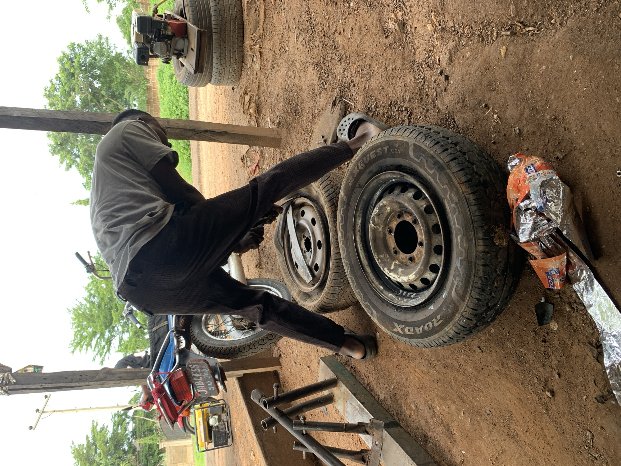 In Gbeleko, Nigeria, a man wearing dark pants and a grey t-shirt is bent over a tire on a dirt ground. He is wearing black clogs. There are several tires stacked nearby, and a generator is on top of two tires in the background. A motorcycle with a blue fuel tank and a red rack is parked behind him. The scene appears to be an outdoor workshop or repair area.